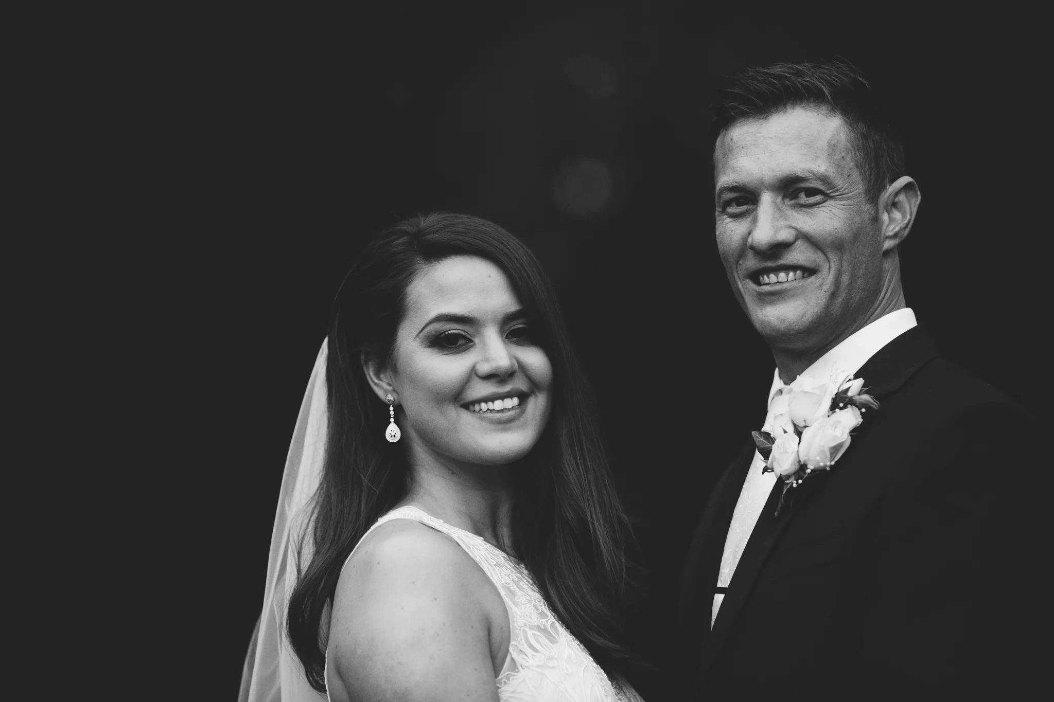Black and white photo of a smiling bride and groom in wedding attire, standing close together outdoors.