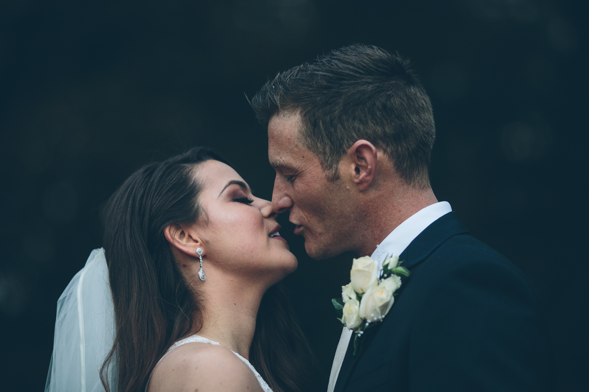 A bride and groom close to each other, about to kiss, outdoors against a dark background.
