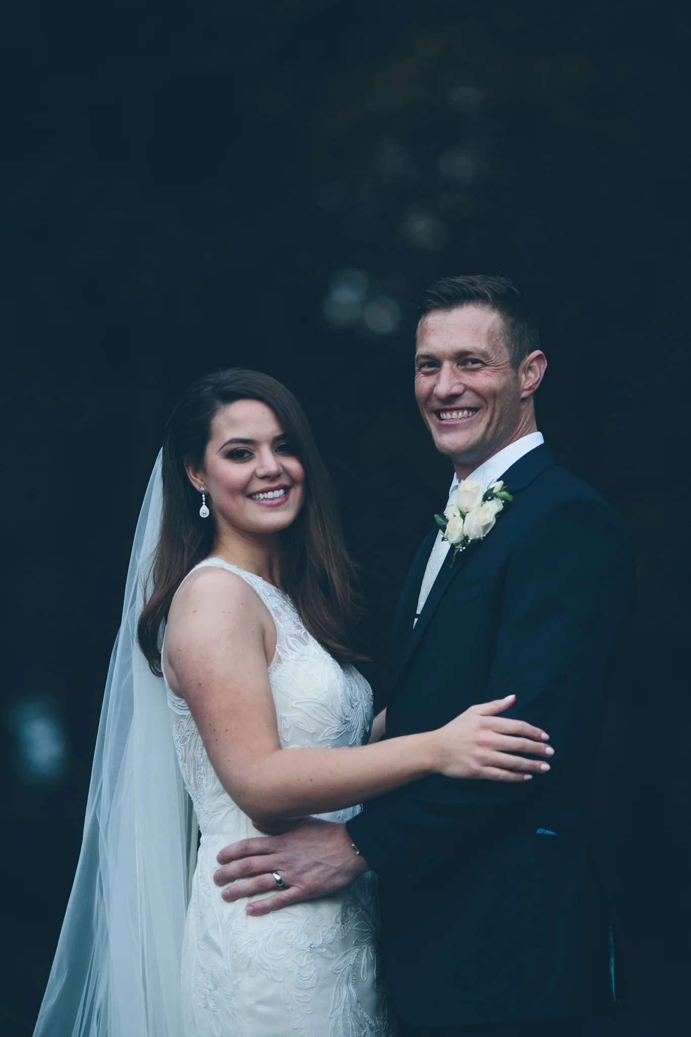 A bride and groom smiling and holding each other outdoors on their wedding day, with a dark, blurred background.