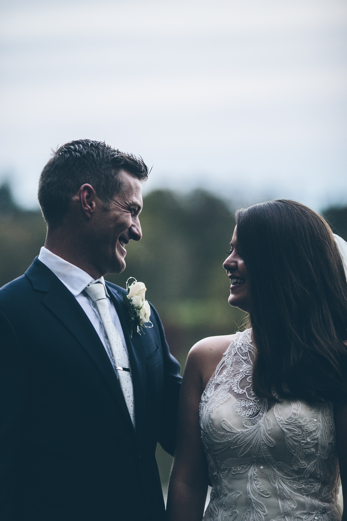 A bride and groom smiling at each other outdoors against a cloudy sky.