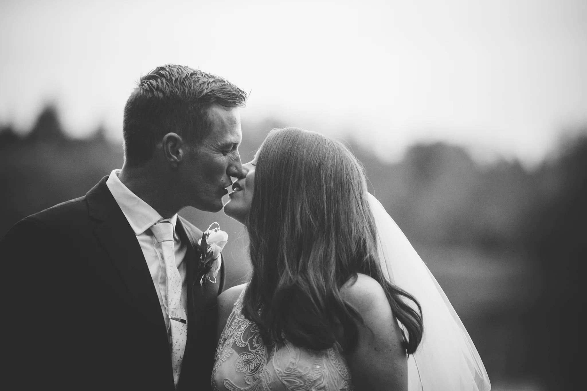 Black and white photo of a bride and groom about to kiss outdoors, with a blurred natural background.