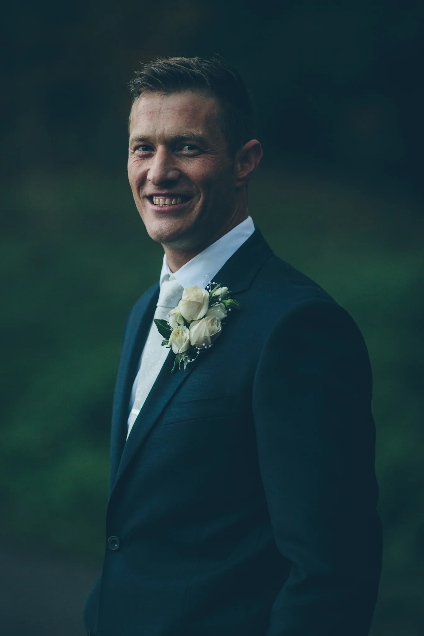 A man in a dark tuxedo with a white shirt and boutonniere, smiling outdoors at dusk.