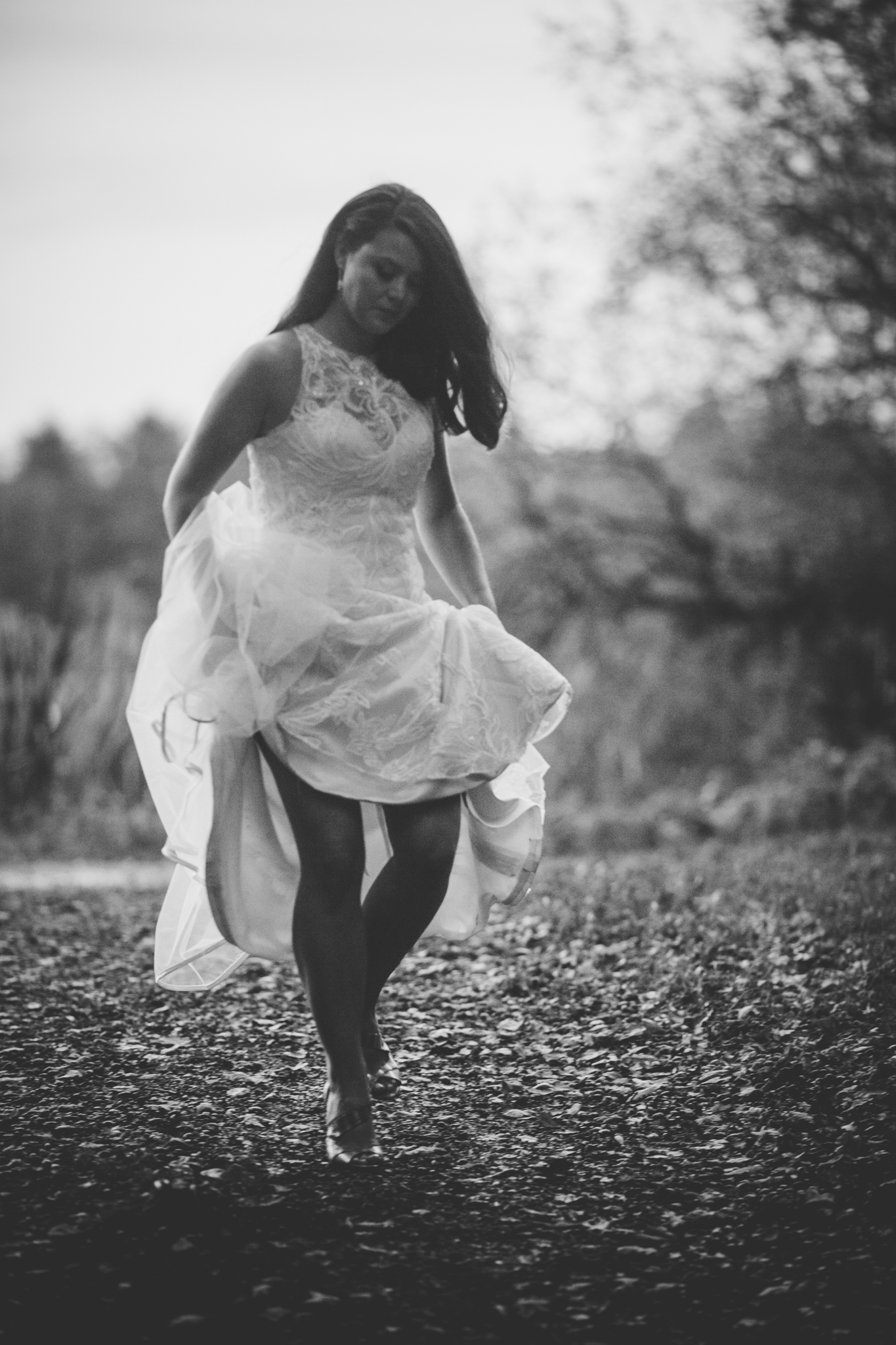 A woman in a white dress walking barefoot on a dirt path outdoors, with trees in the background, captured in black and white.