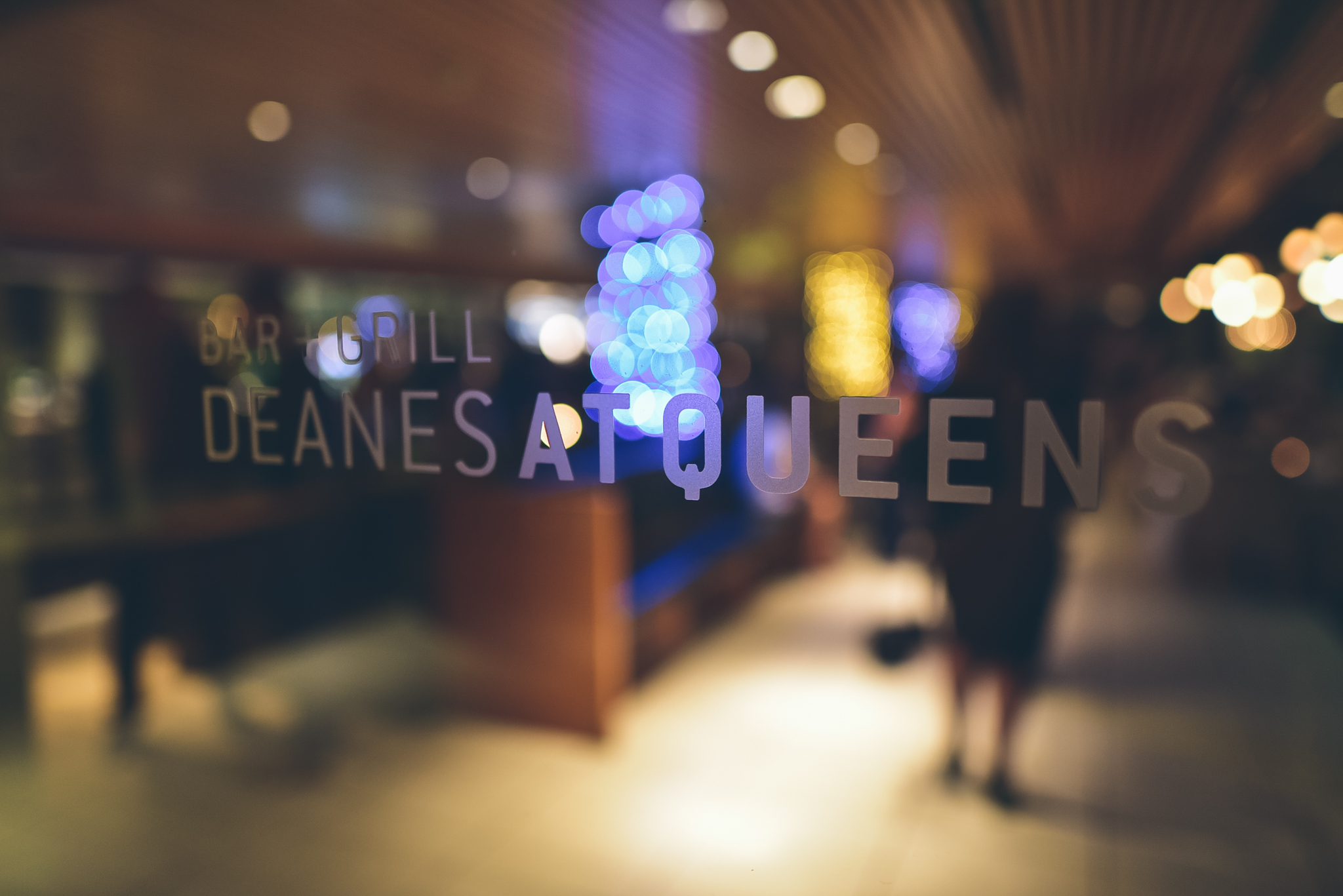 Blurred view of a bar or restaurant with neon and string lights, seen through a window with the text 'Deanes at Queens' on it.