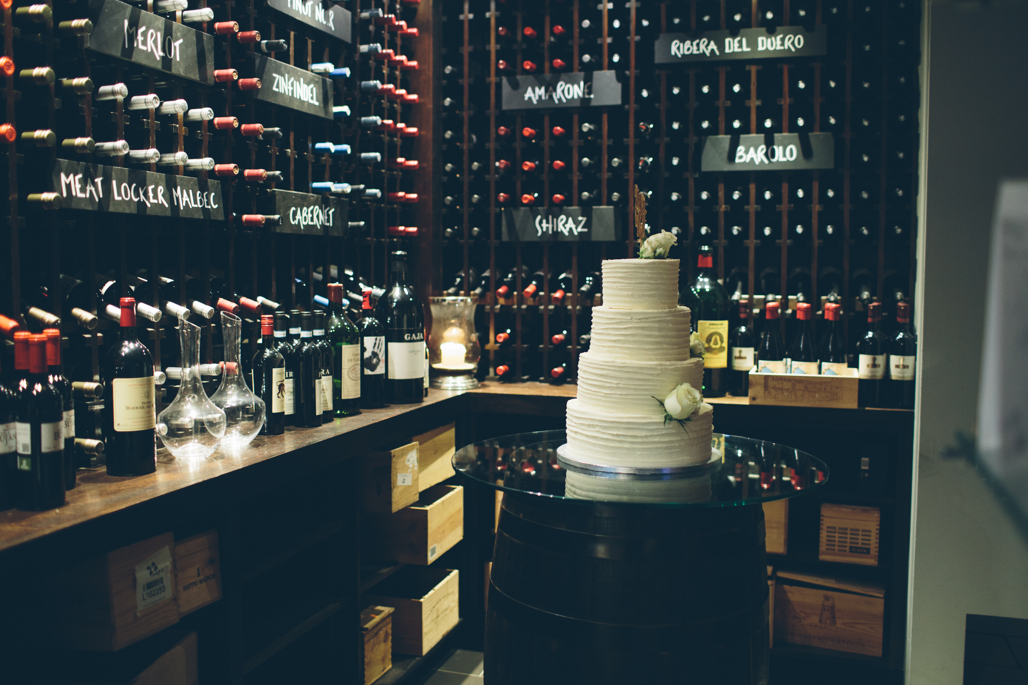 A white wedding cake with flowers on top on a black barrel in a wine cellar, with bottles of wine and labeled wine racks in the background.