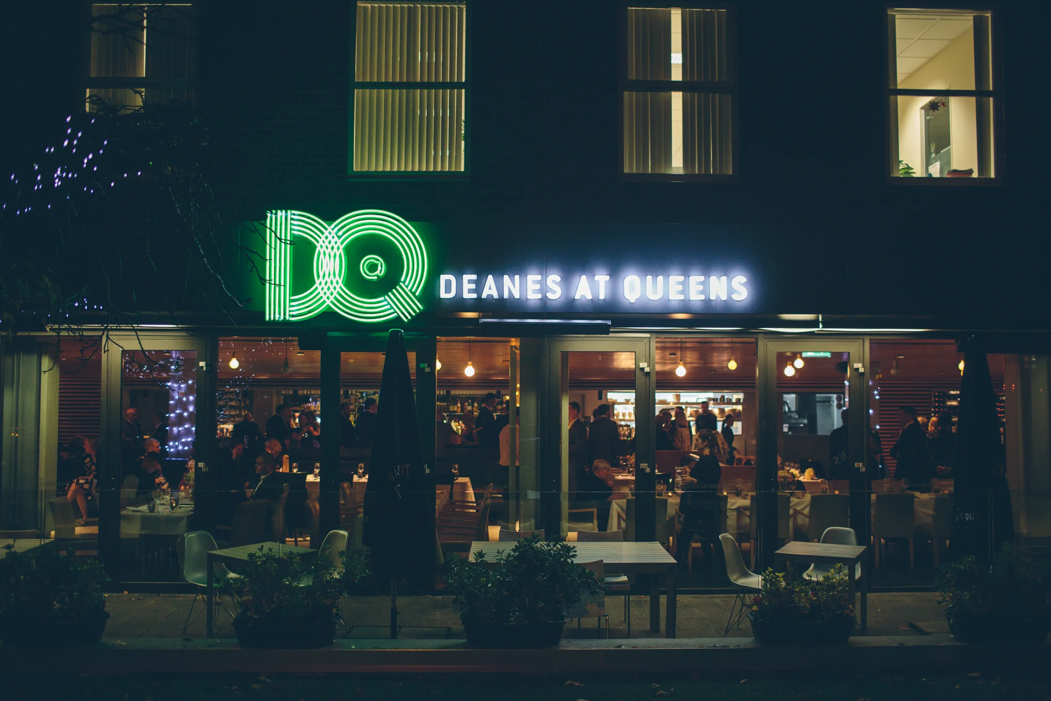 Night view of a restaurant named 'Deanes at Queens' with illuminated signage, showing people dining inside through large glass windows, decorated with string lights and outdoor seating in front.