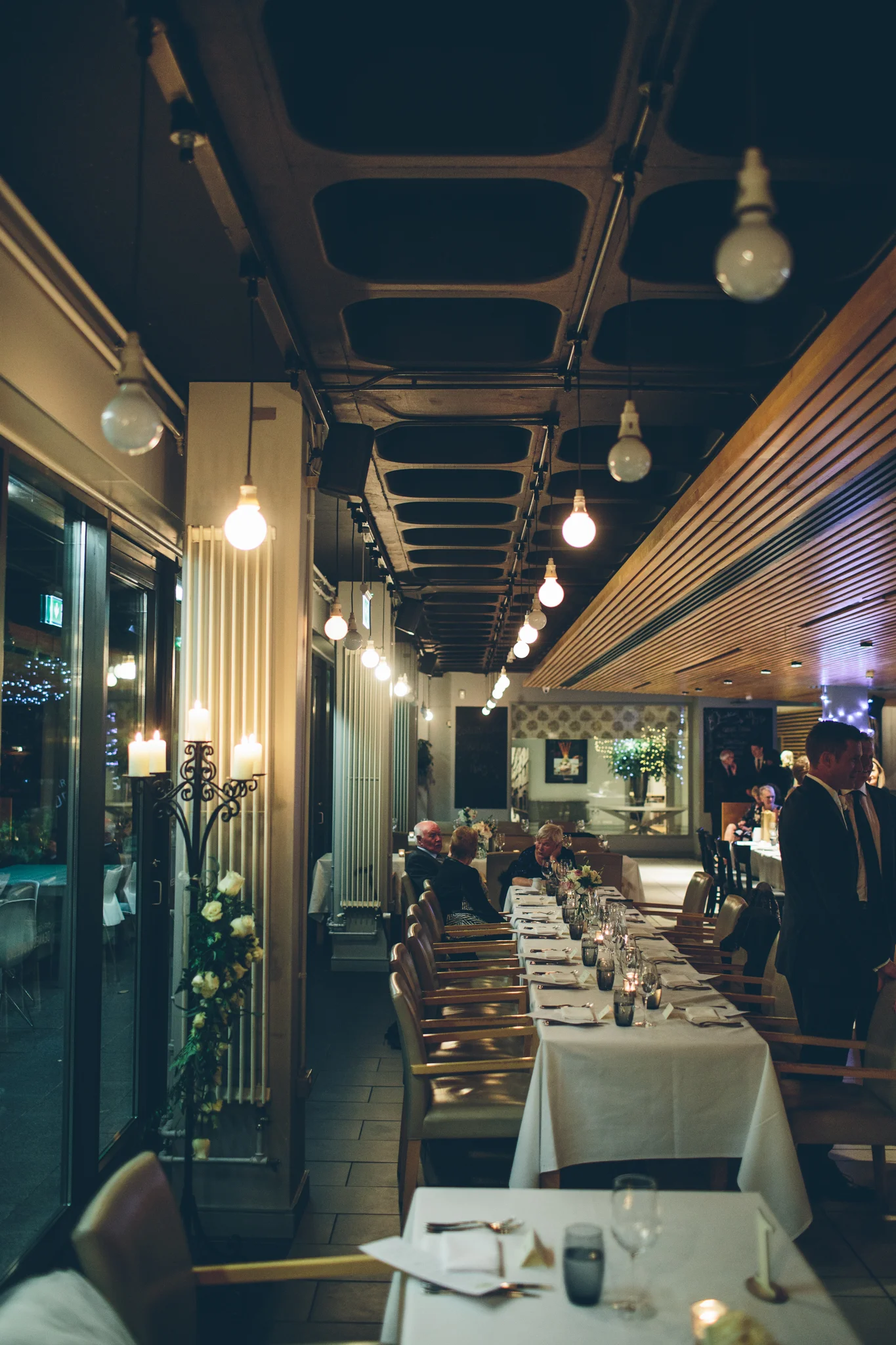 Interior of a restaurant with decorated tables and ambient lighting, some guests seated and some standing.