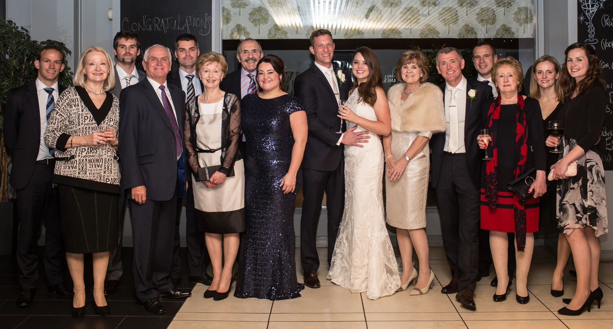 Group of people dressed in formal attire at a celebration, with wedding couple in center, inside a decorated venue.