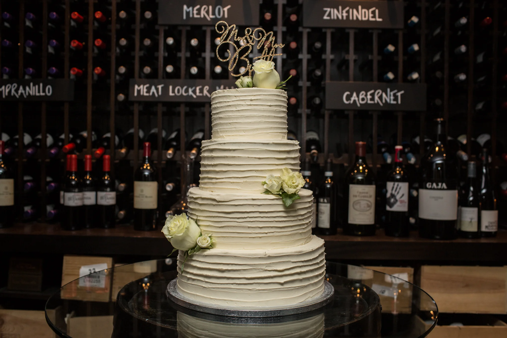 Three-tier white wedding cake with white roses and a gold "Mr & Mrs" topper in a wine cellar setting.