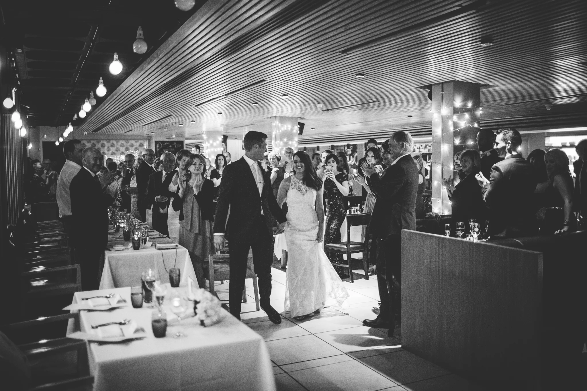 Black and white photo of a wedding reception with the bride and groom walking hand-in-hand through a crowd of guests in a decorated reception hall.
