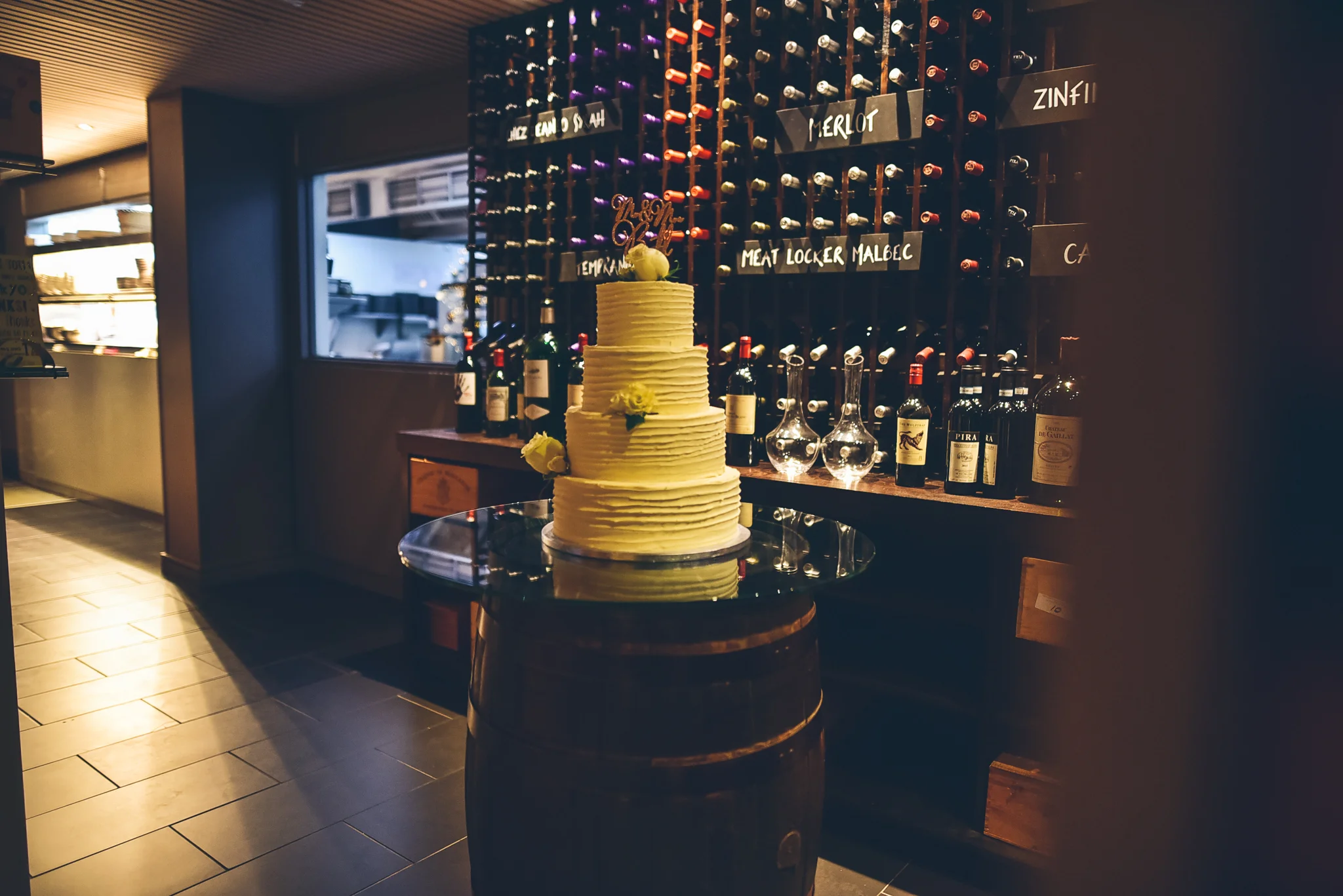 A three-tier white cake with yellow flowers and a 'Happy Birthday' topper on a glass top on a barrel, in front of a wine wall with bottles and wine glasses in a dimly lit restaurant or winery.