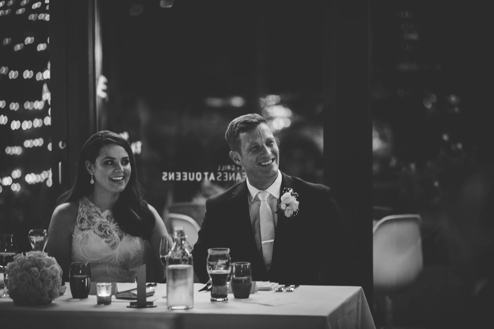 A black and white photo of a newlywed couple seated at a wedding reception table, smiling and looking in opposite directions. The bride is wearing a lace wedding dress with pearl earrings and has long dark hair. The groom is wearing a dark suit with 