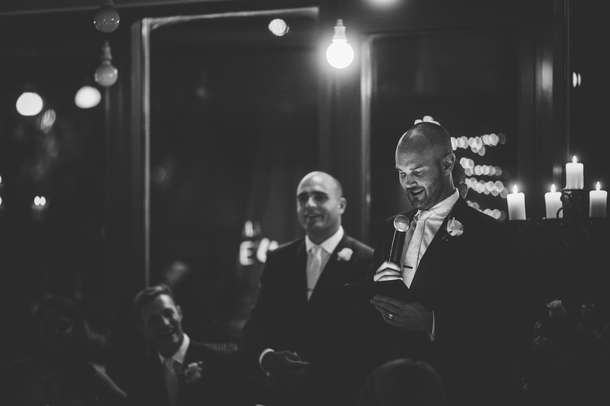 Groom giving a speech at his wedding reception, holding a microphone and reading from a paper, with wedding guests in the background, candles on the table, and soft decorative lighting.