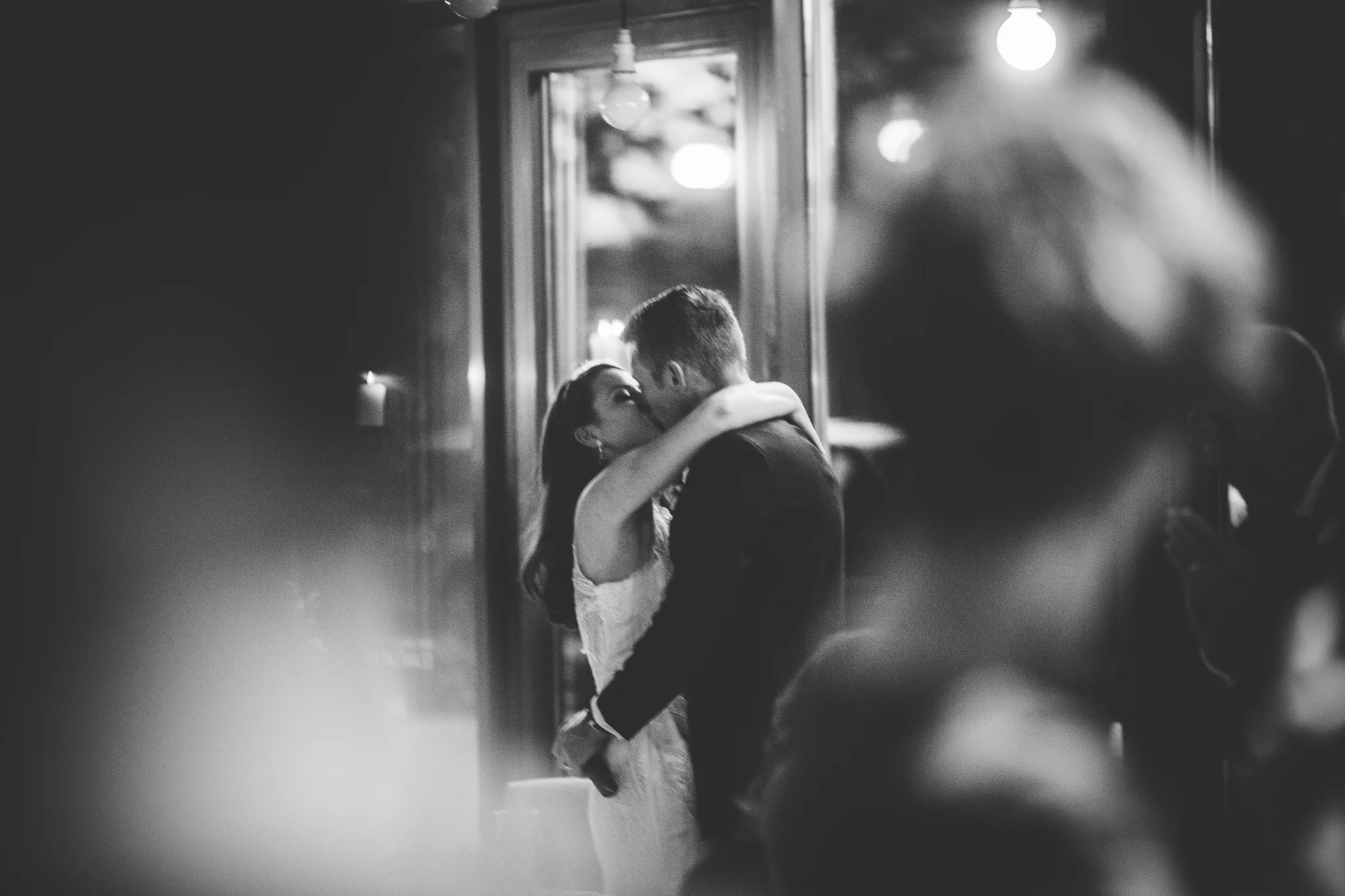 A black and white photo of a couple kissing, with the woman wearing a sleeveless dress and the man in a suit, in an intimate moment at a social event.