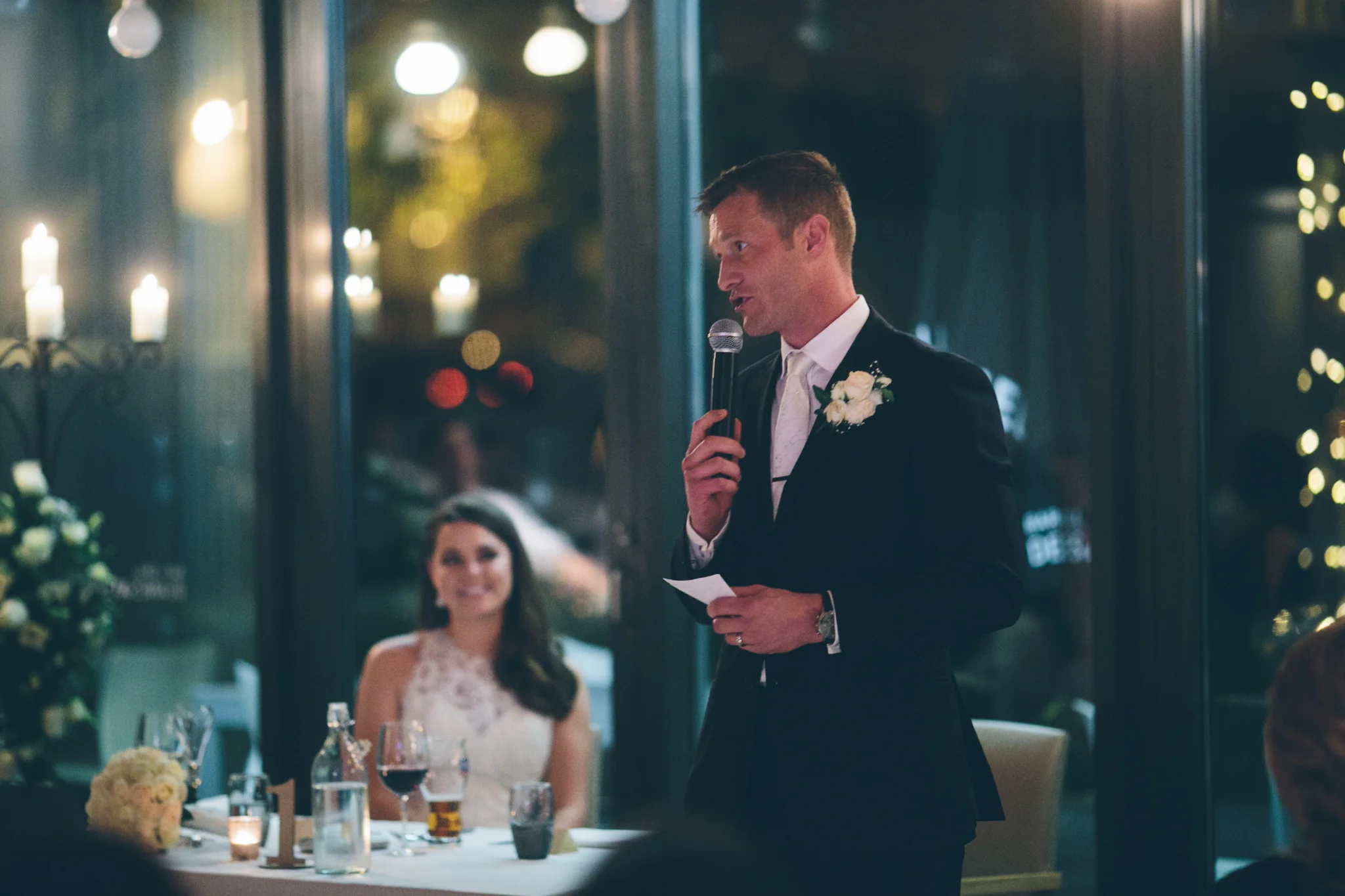 A man in a tuxedo giving a speech at a wedding reception, holding a microphone and a piece of paper, with a woman in a wedding dress smiling in the background.