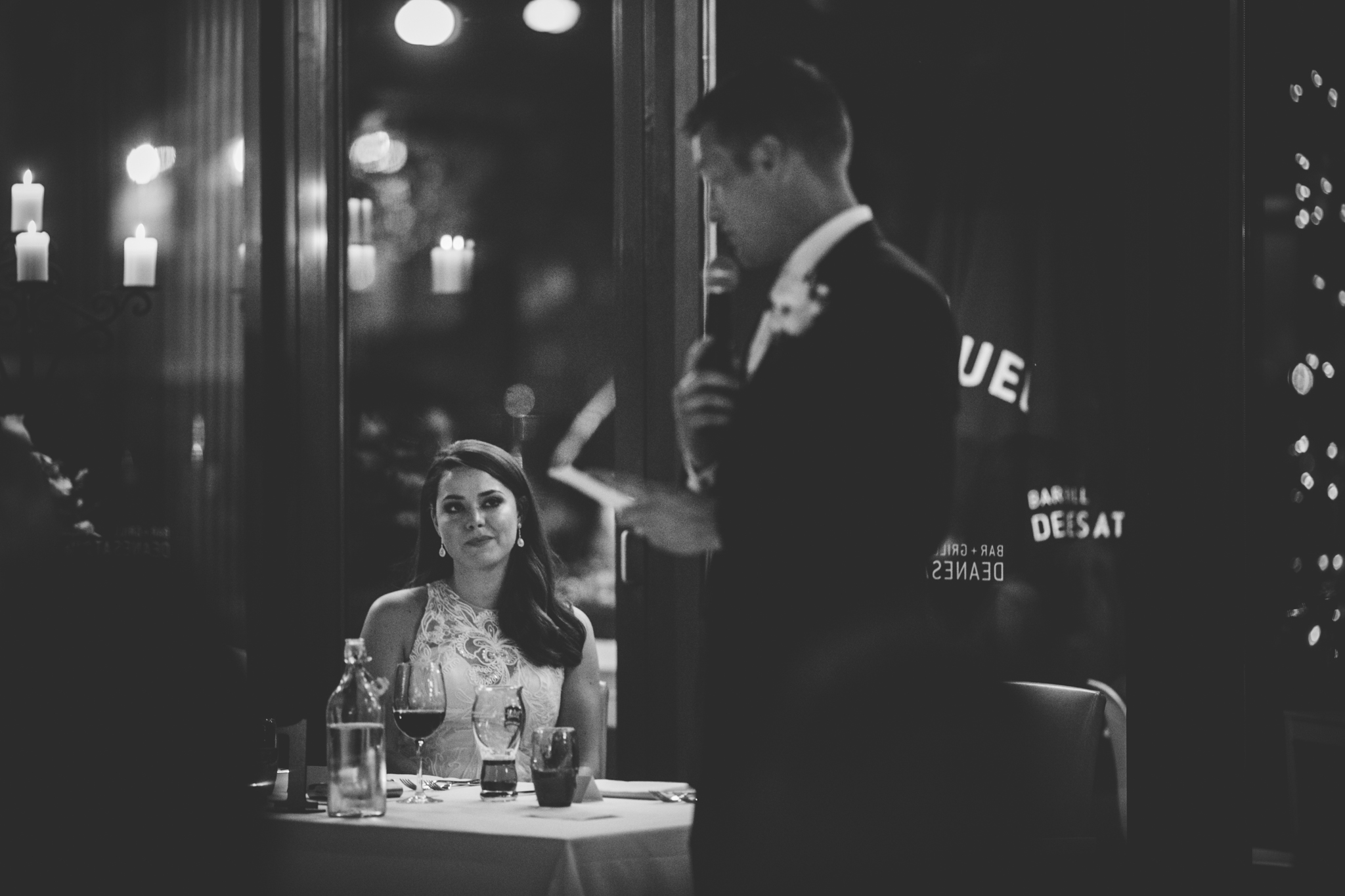 A woman in a white dress sitting at a table in a restaurant, looking up, with a man in a suit standing nearby reading a menu. The scene is in black and white, and the table is set with wine glasses and bottles.