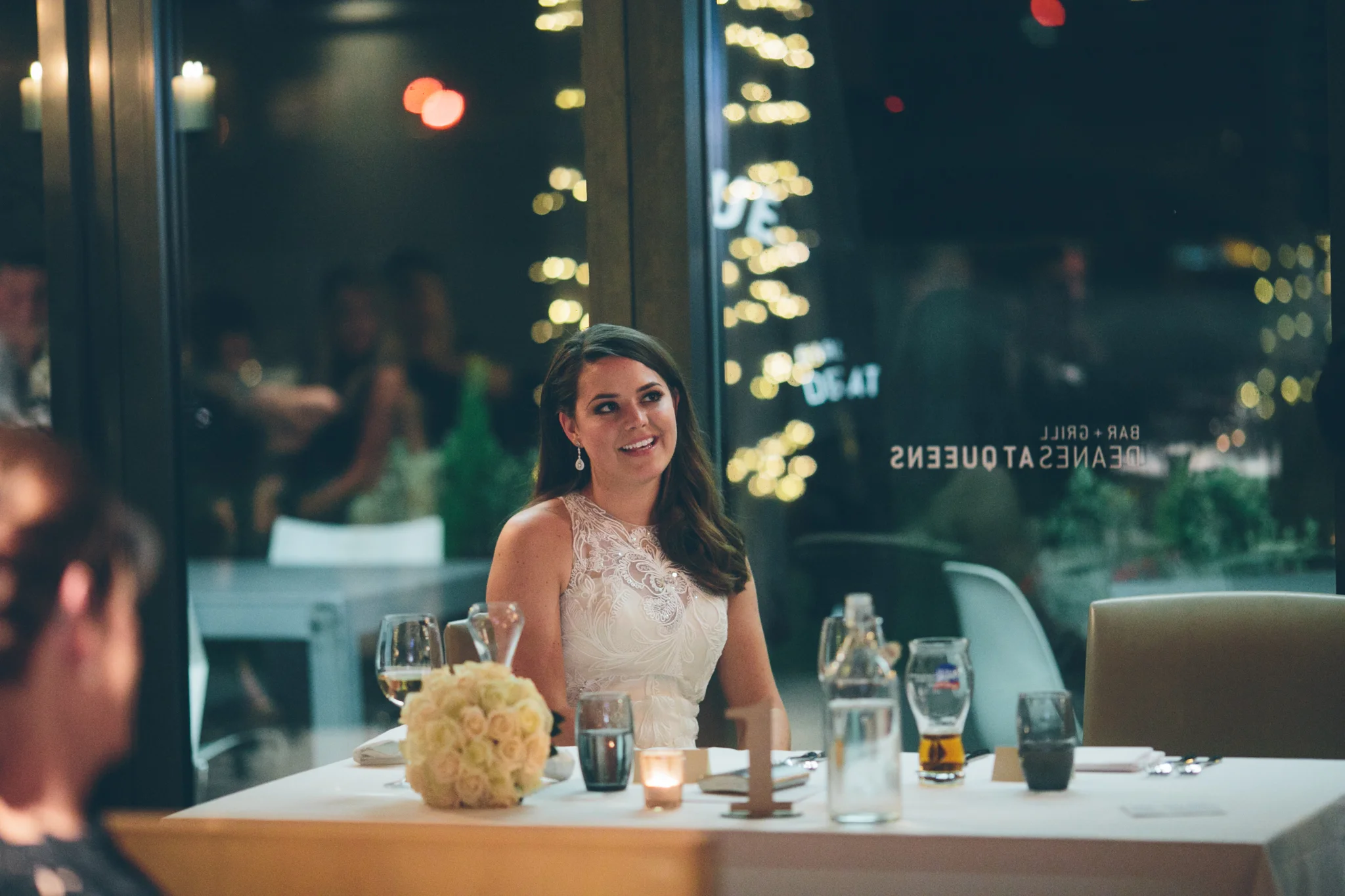 A young woman in a white dress smiling at a dinner party, sitting at a table with drinks and a floral centerpiece, inside a glass-walled restaurant at night.