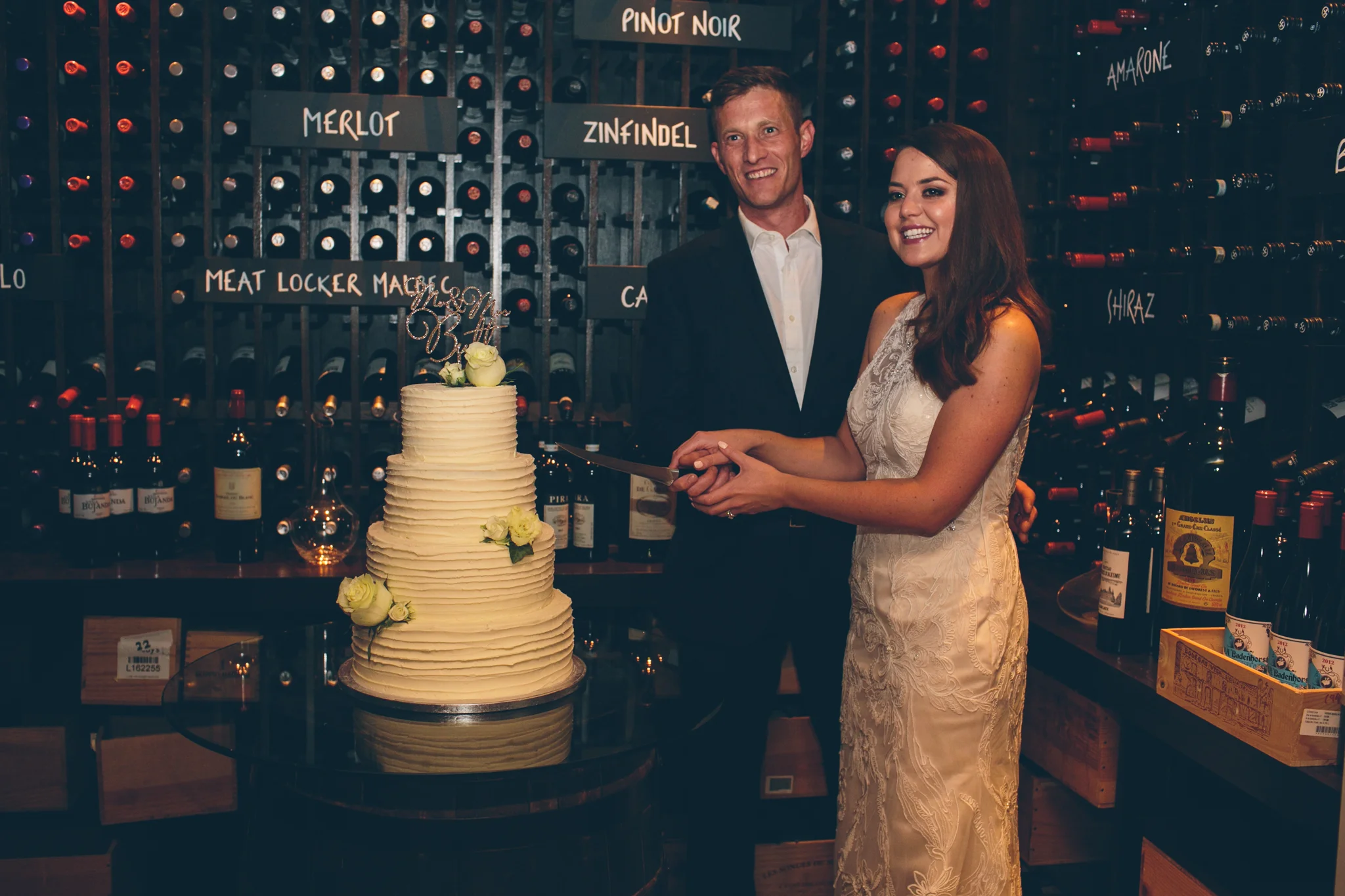A woman in a light-colored lace dress and a man in a dark suit hold a cake knife together, smiling at a wedding celebration with a three-tiered cream-colored wedding cake decorated with white roses. The background features a wine cellar with bottles 