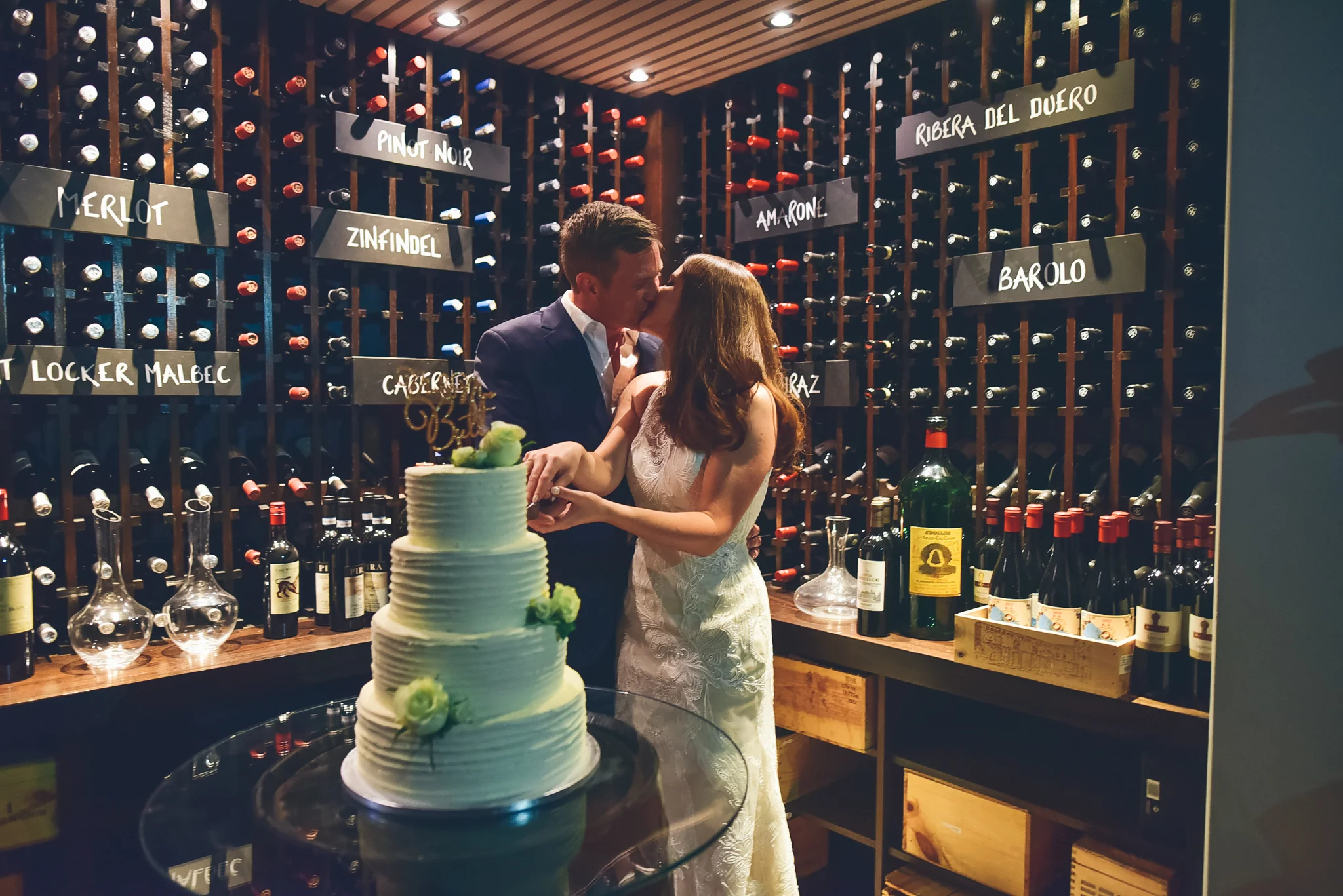 A couple kisses at their wedding reception in a wine cellar, with a wedding cake in the foreground and bottles of wine on shelves behind them.