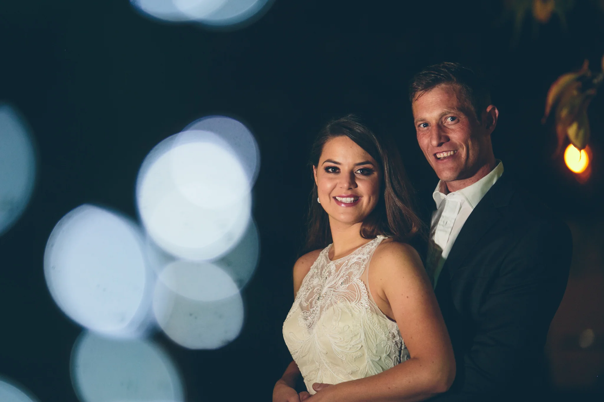 A smiling couple dressed in formal attire at night, with prominent bokeh light effects in the foreground.