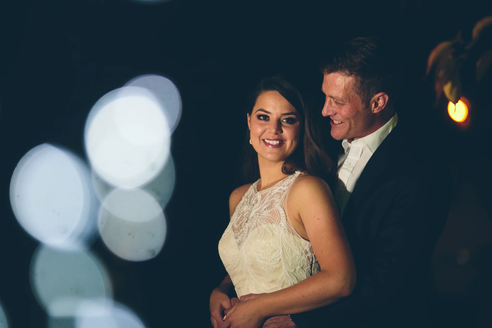 A smiling woman in a white dress with lace detail and a man in a black suit, both standing close together at night with blurry bright lights in the background.