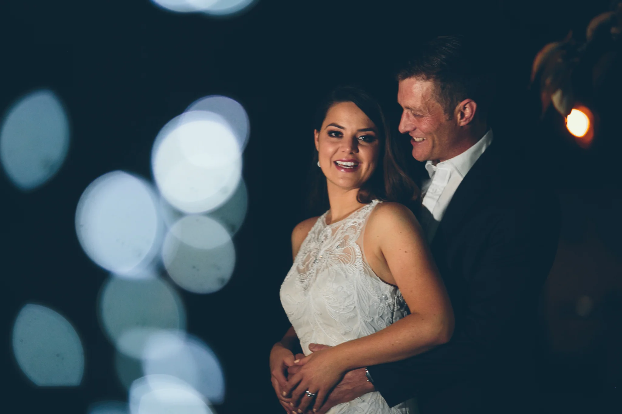 A couple at a nighttime event, the woman in a white lace dress and the man in a dark suit, smiling and embracing each other with blurred bokeh lights in the background.