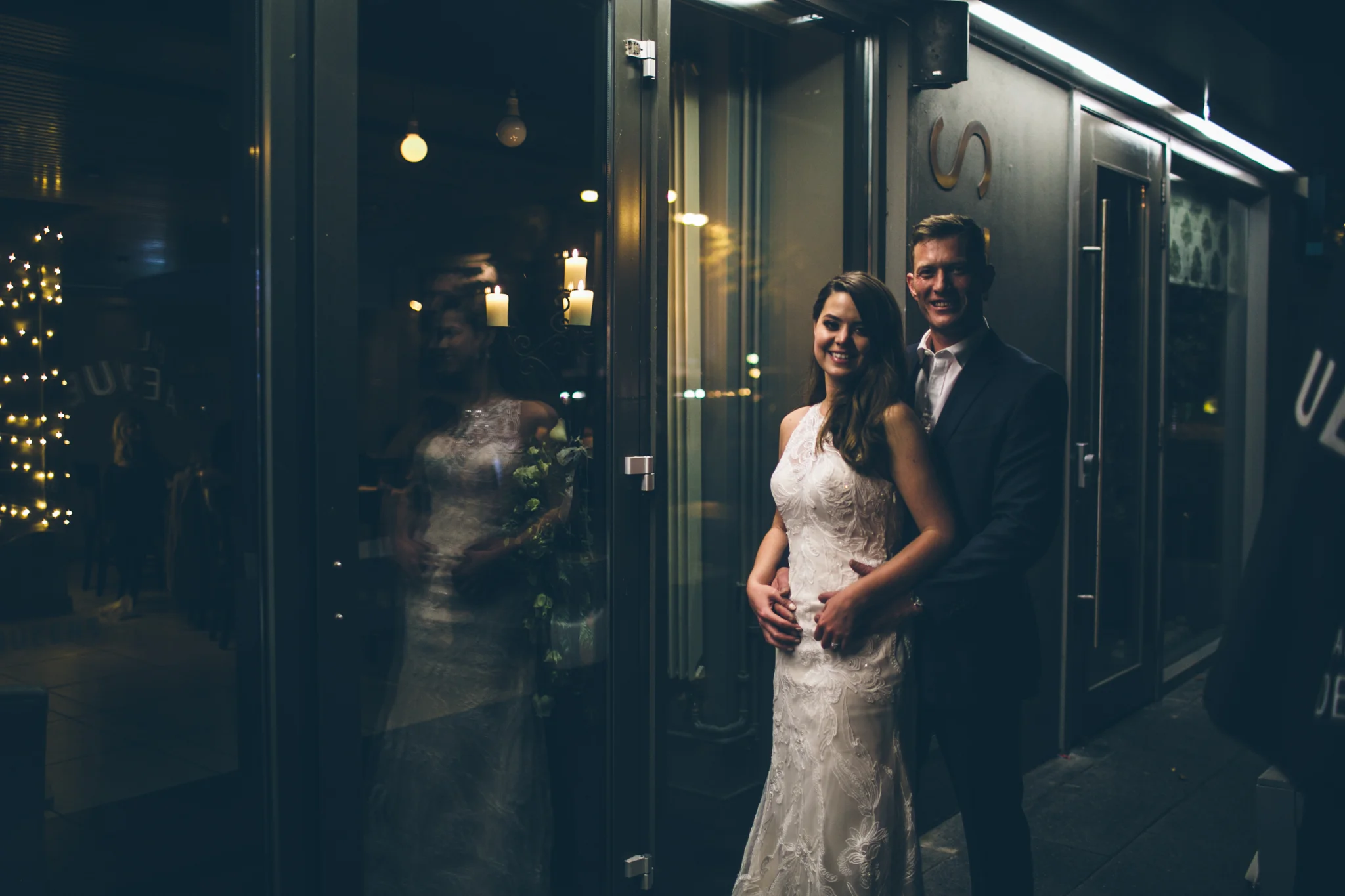 Couple dressed in wedding attire standing outside a building at night, with the woman holding a bouquet and smiling at the camera.