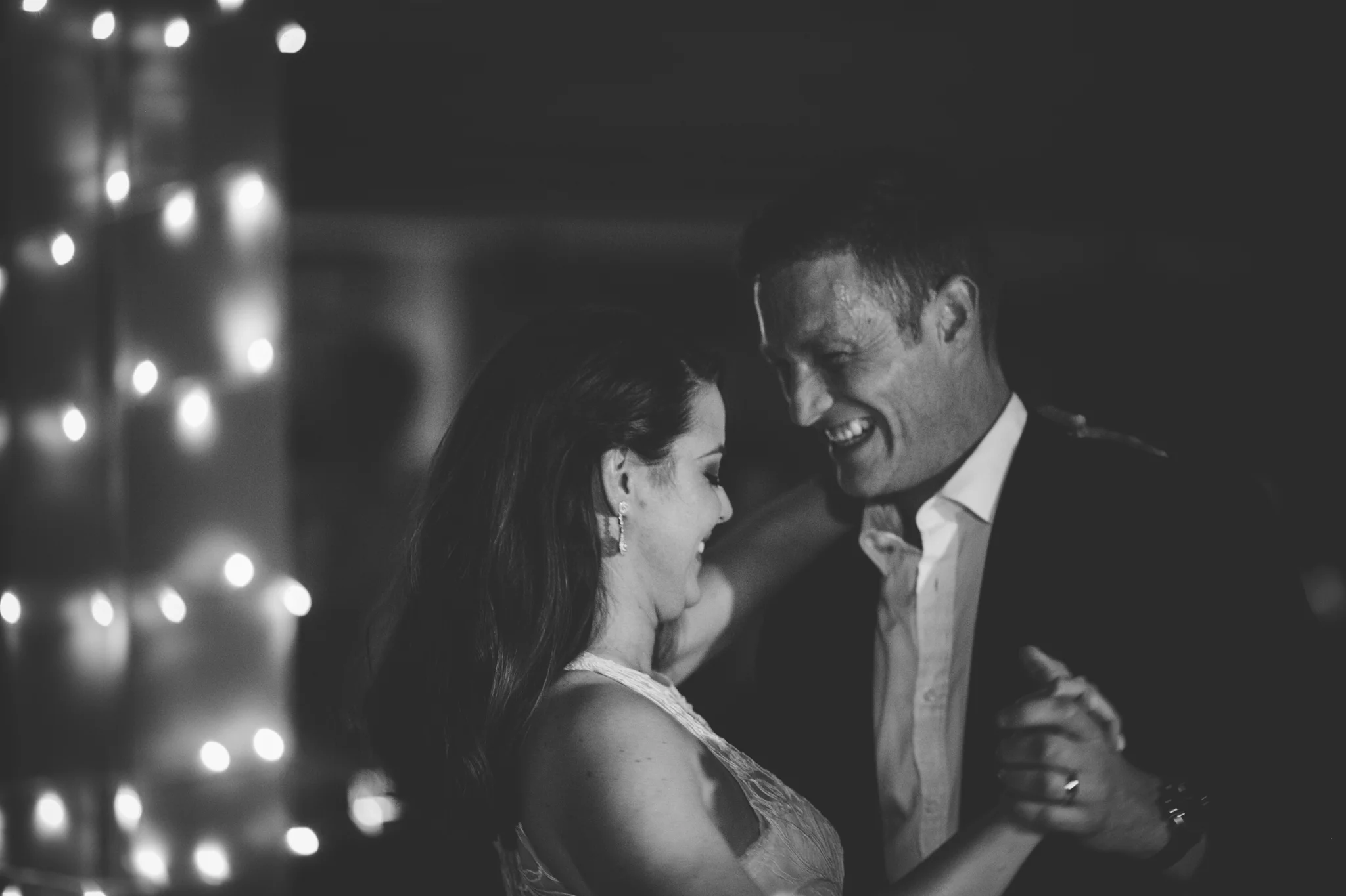 A black and white photo of a smiling couple dancing closely together, illuminated by soft lighting and fairy lights in the background.