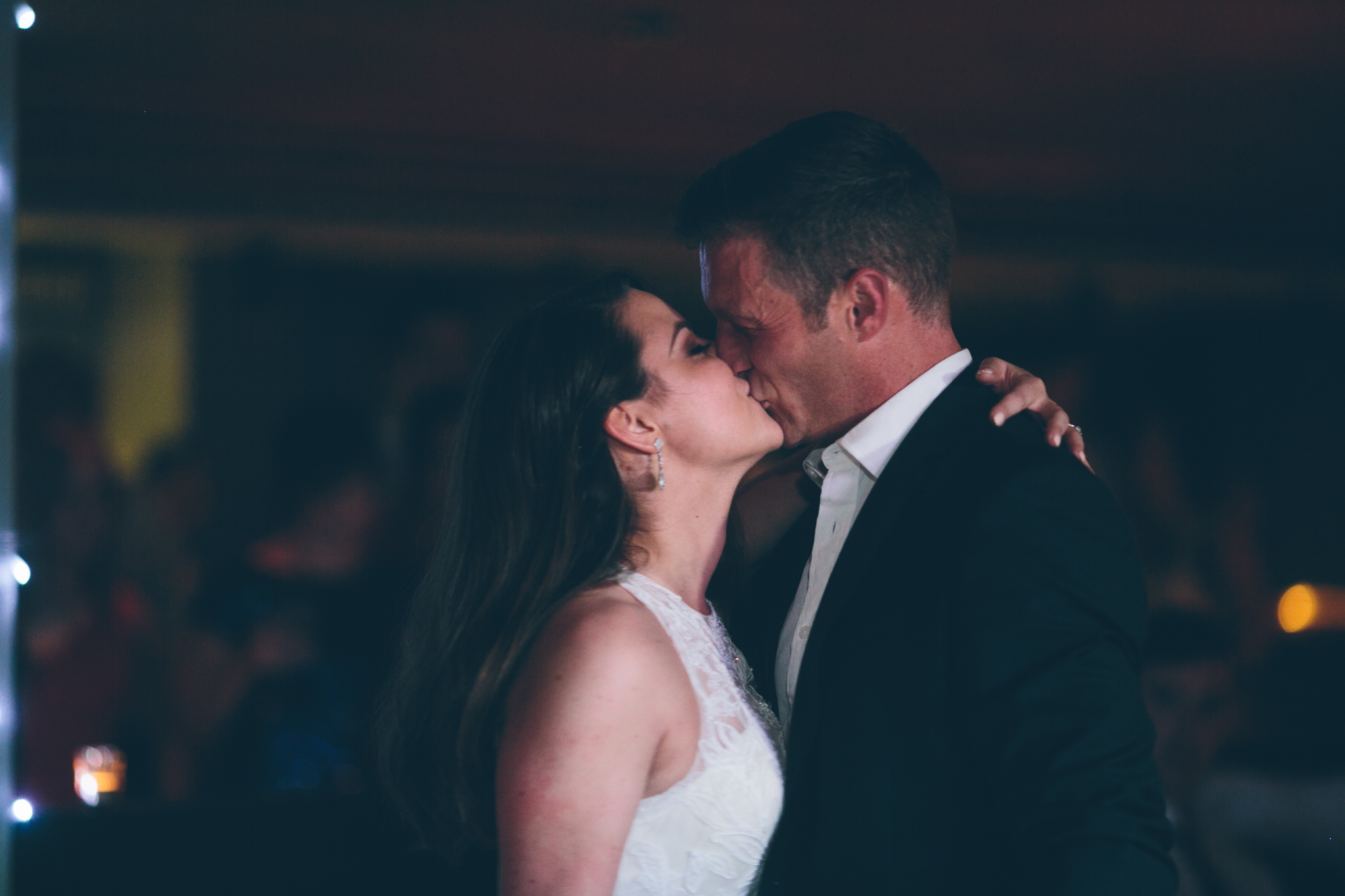 A couple kissing at a wedding reception, with the woman in a white laced dress and the man in a black suit and white shirt, in a dimly lit venue.
