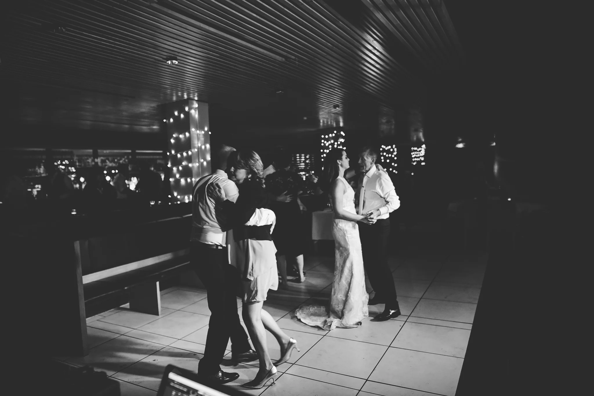 Black and white photo of couples dancing and embracing at a wedding reception, with string lights and a bar in the background.