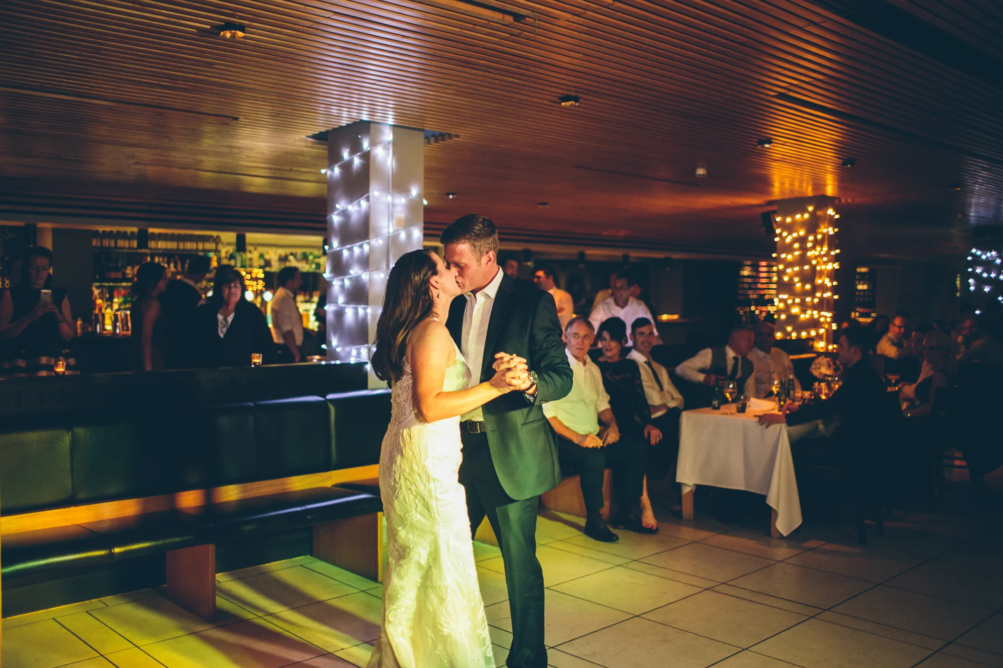 A bride and groom are dancing closely in a dimly lit reception hall with friends watching in the background, decorated with string lights on columns.