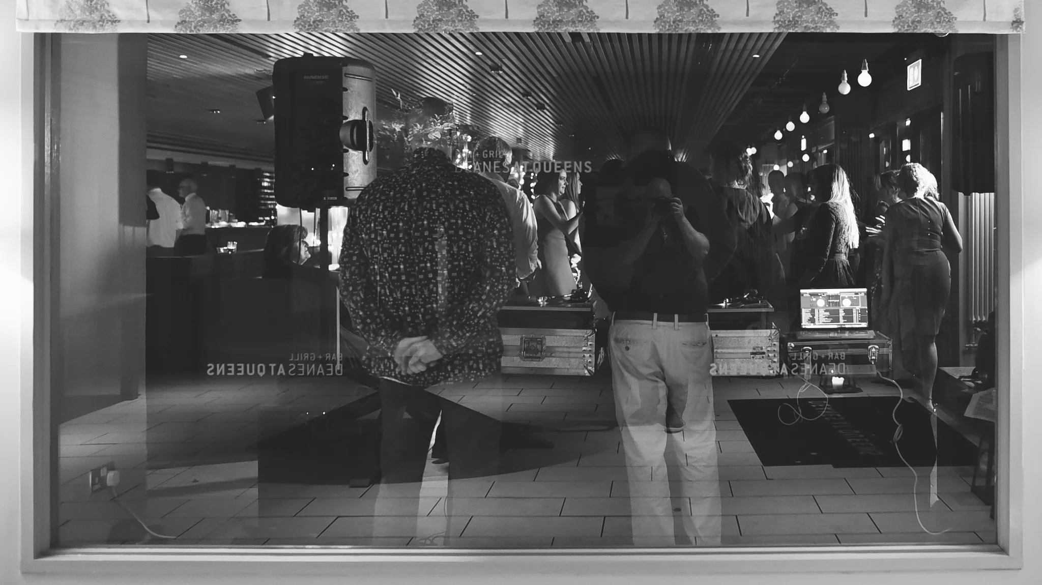 View through glass window of a dance party at an indoor venue with people dancing, DJ equipment, and a speaker, reflected with the photographer taking a picture.