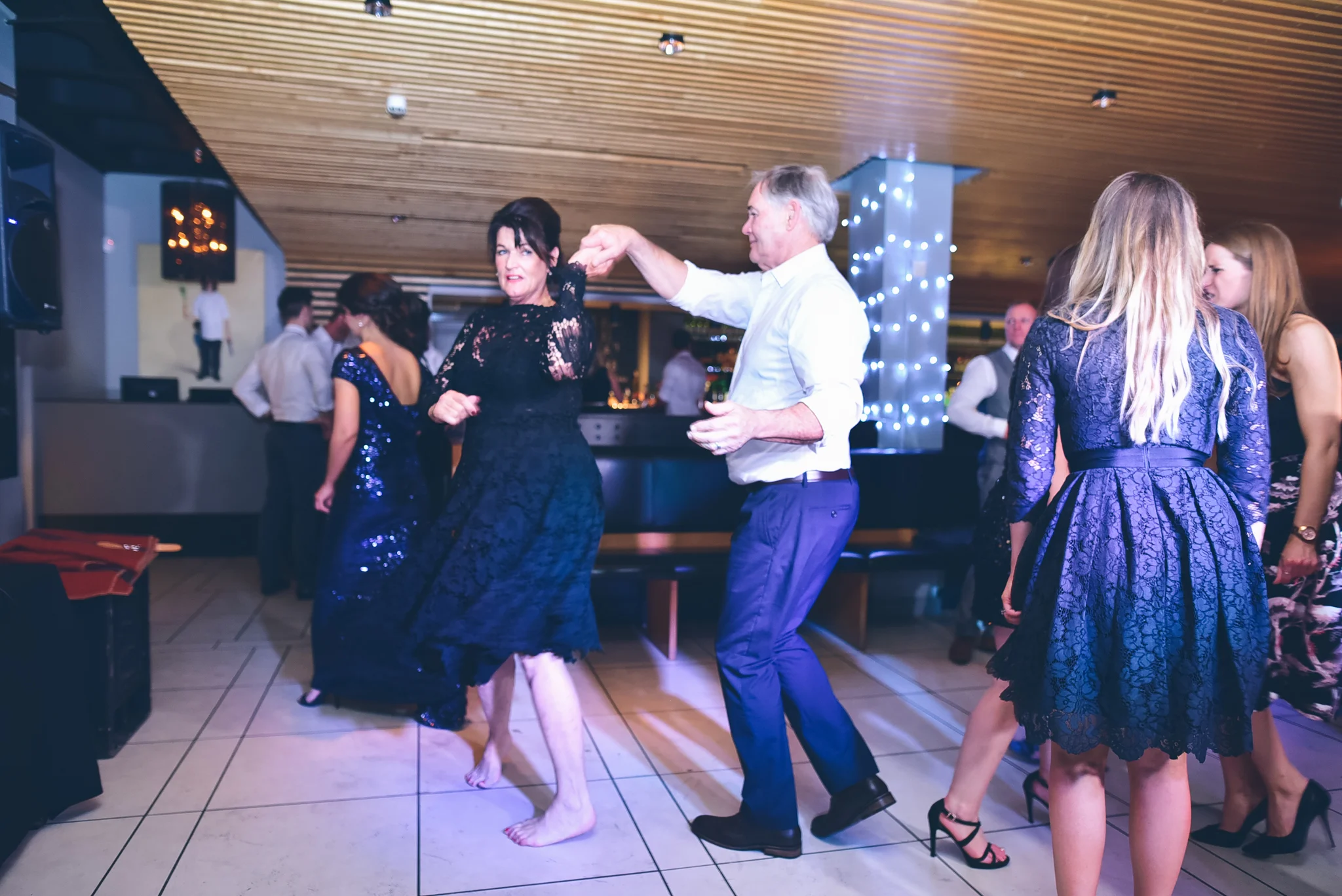 People dancing at a party or celebration in an indoor venue, with wooden ceiling and festive lighting.