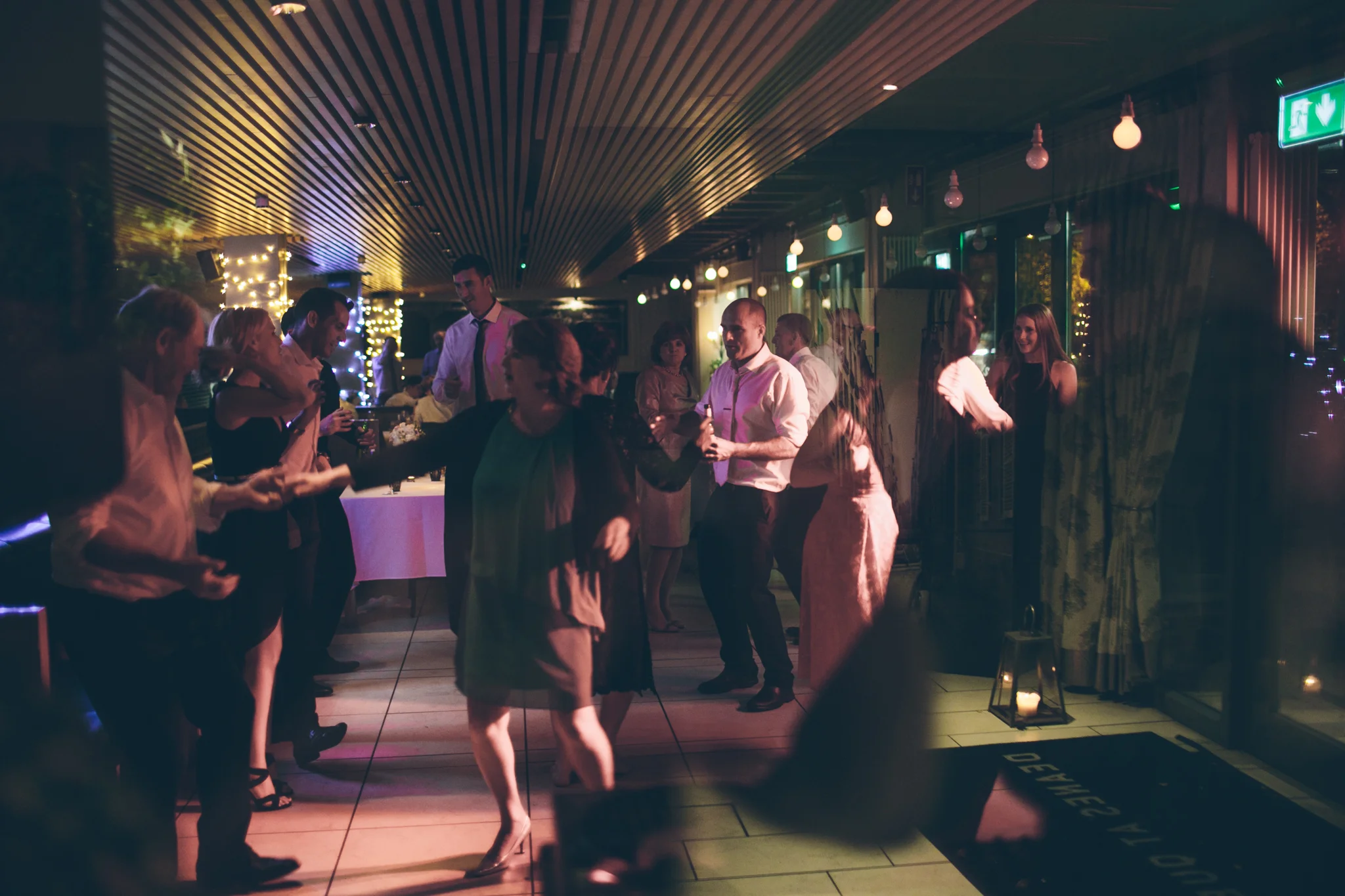 People dancing and socializing at an indoor party or celebration with dim lighting and decorative string lights.