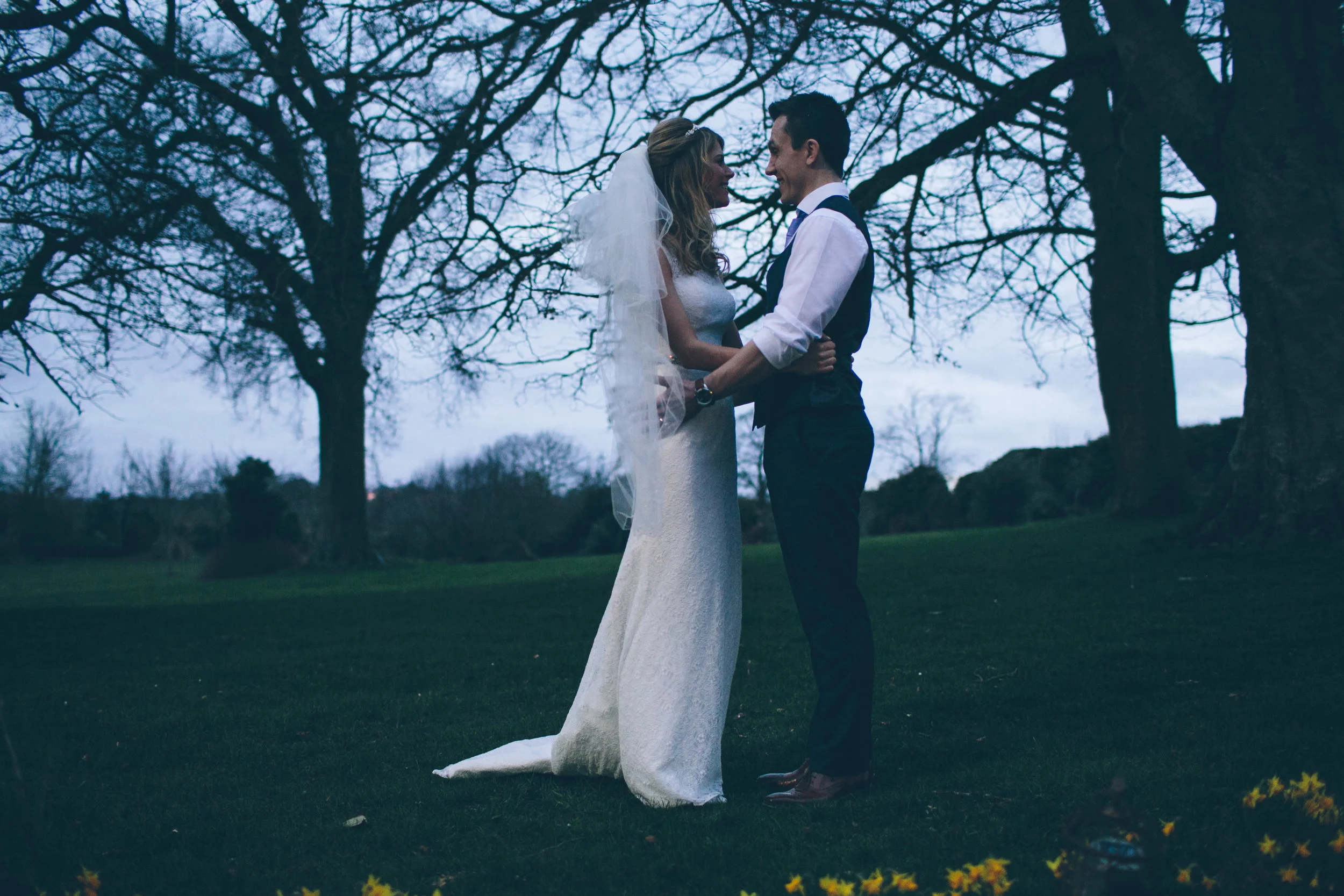 A bride and groom standing close together outdoors on a grassy area surrounded by trees, sharing an intimate moment on their wedding day.