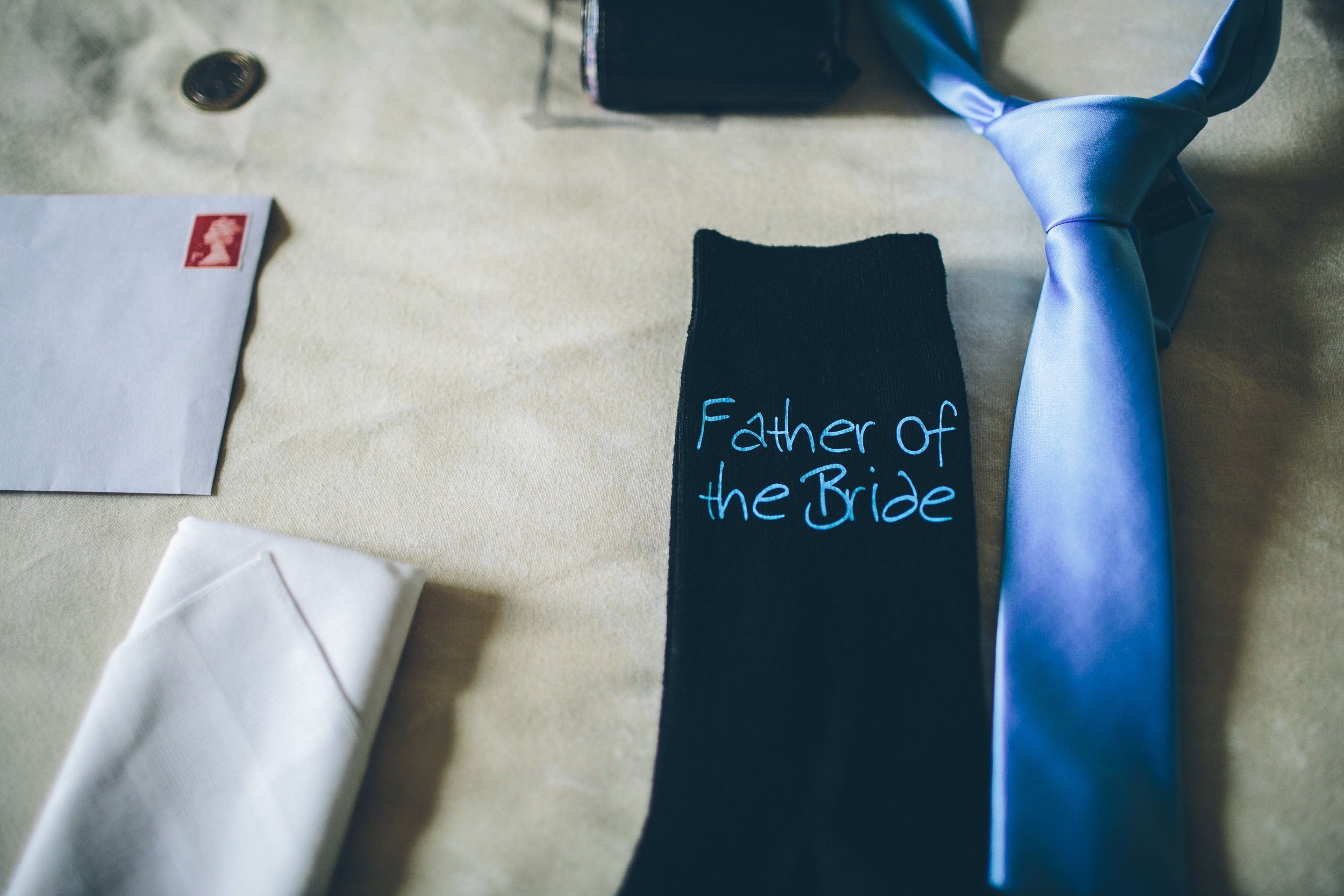 A dark sock with blue embroidery that reads 'Father of the Bride' placed on a table next to a light blue satin tie and a white cloth napkin, with an envelope and a small perfume bottle visible in the background.