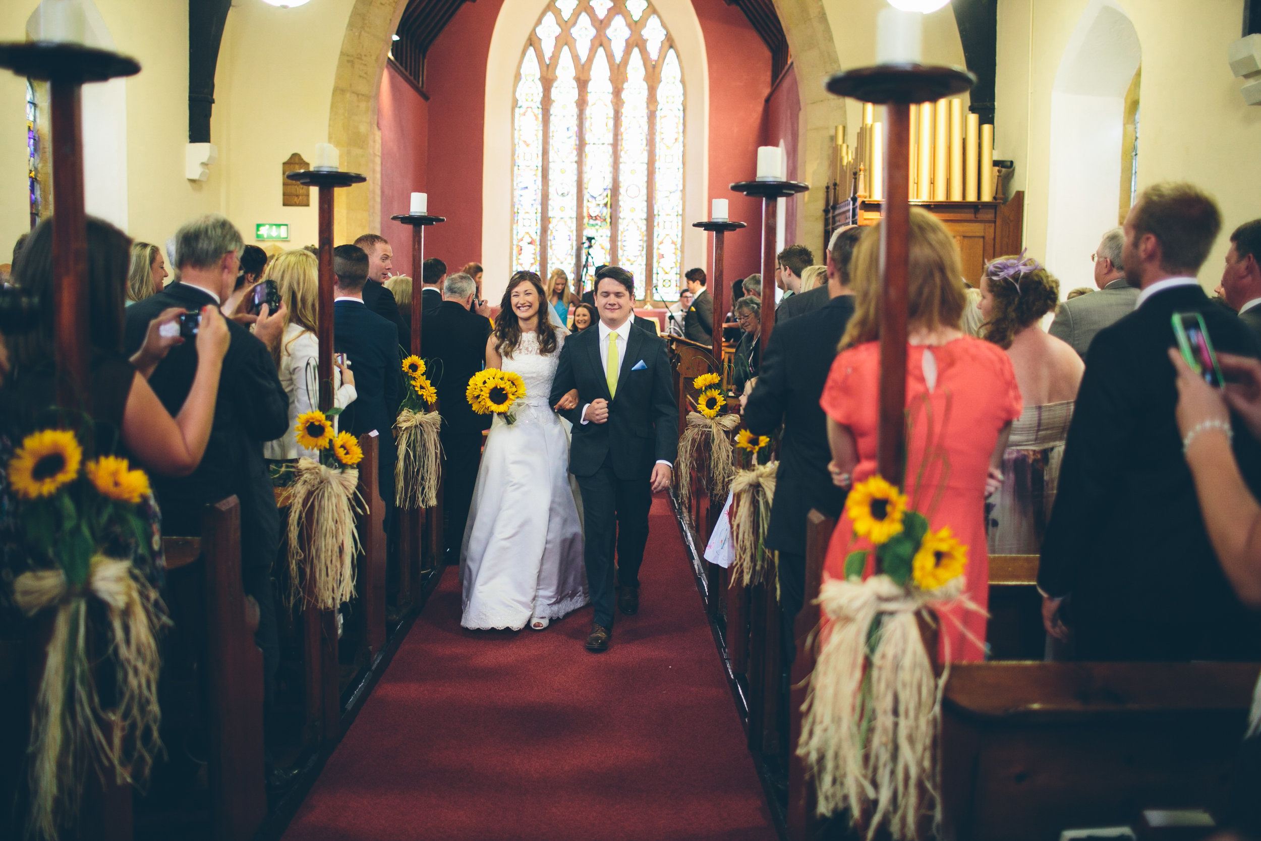 A bride and groom walking down the aisle inside a church during their wedding ceremony, surrounded by guests taking photos and watching.