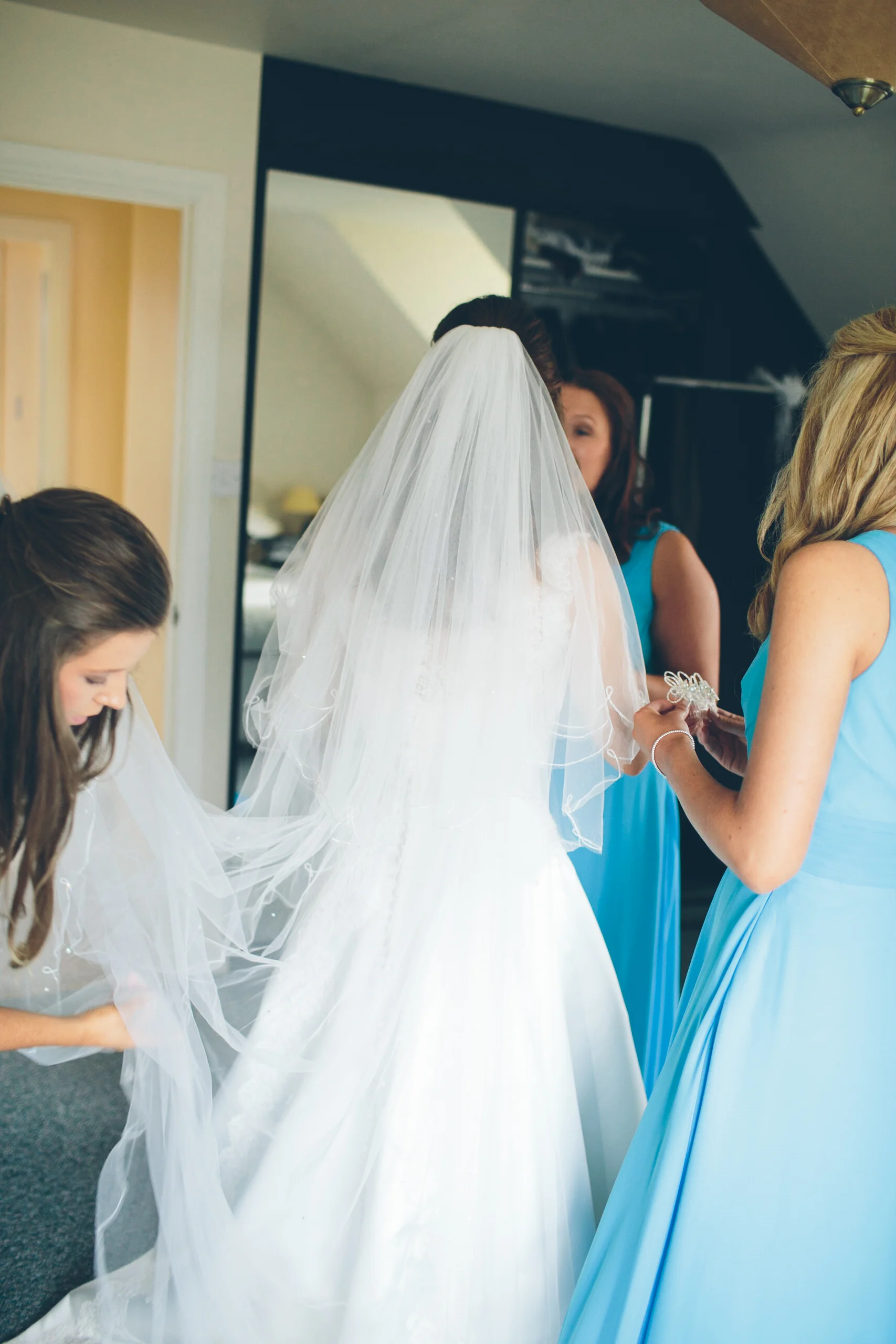 A bride in a white wedding gown and veil is being assisted by women in blue dresses, possibly bridesmaids, in a room preparing for a wedding.