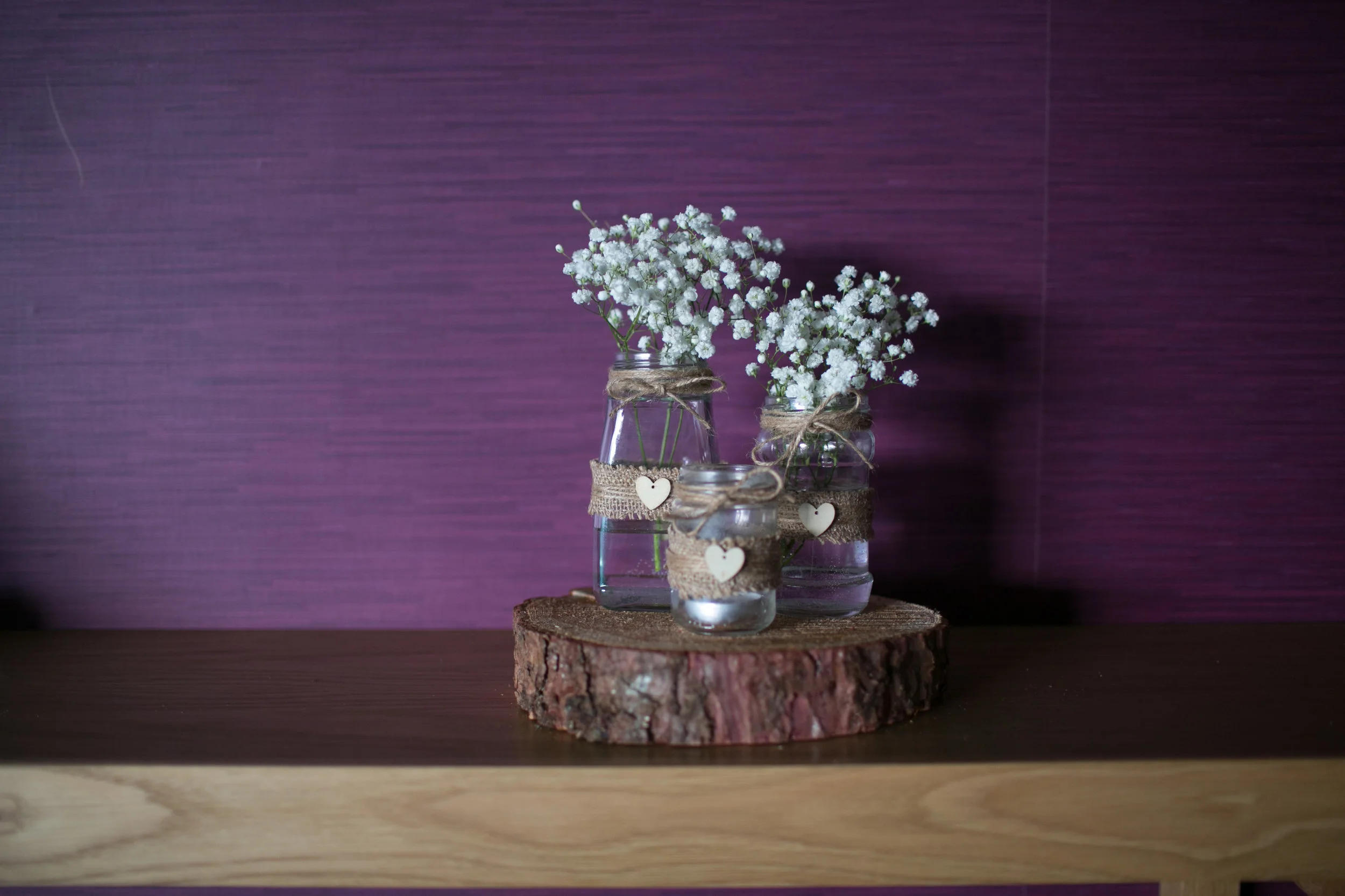 Three glass jars containing white baby's breath flowers, decorated with burlap and twine, placed on a wooden slab against a purple wall background.