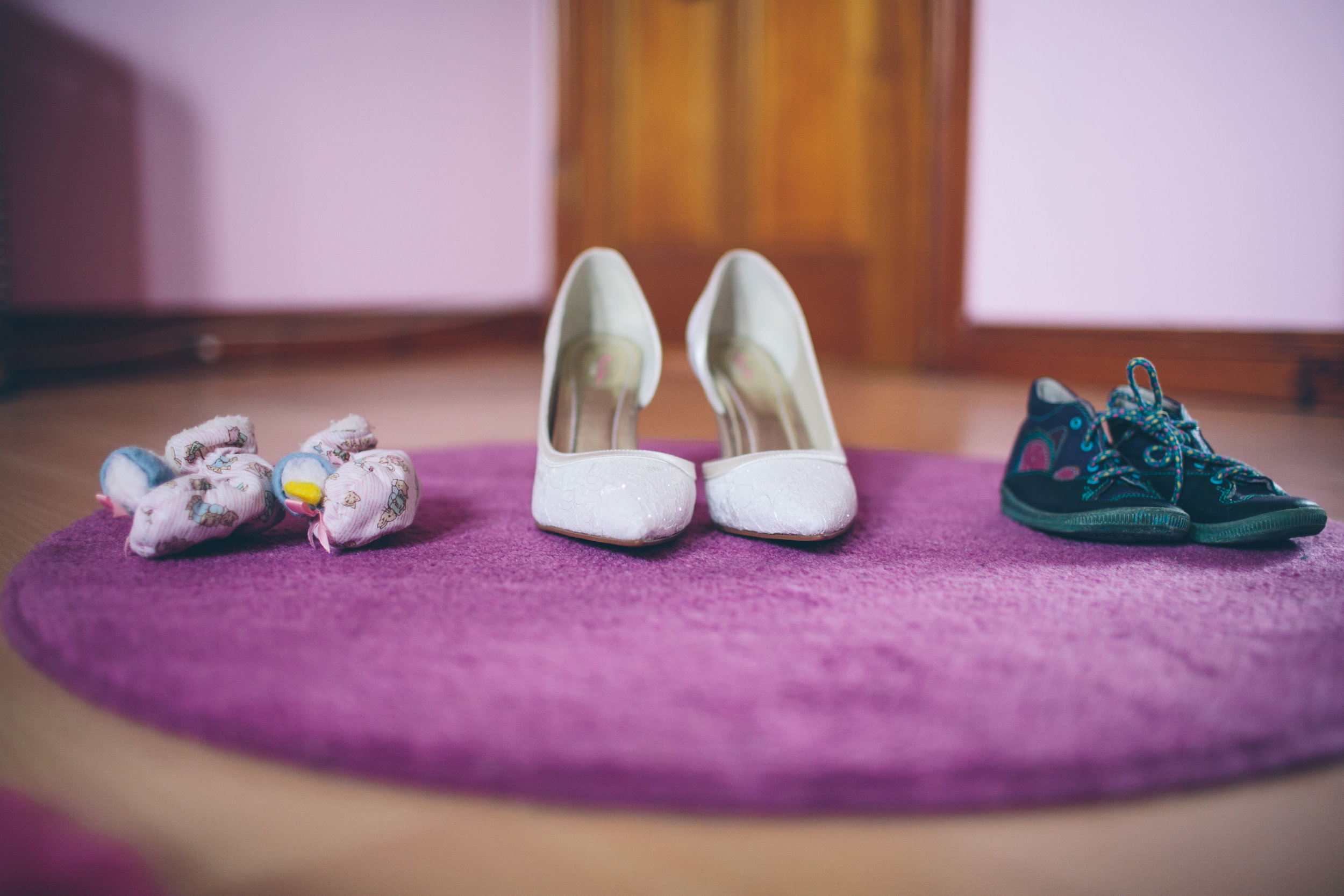 Pair of white high-heeled shoes in the center, pink baby socks on the left, and black sneakers on the right, all placed on a purple circular rug.