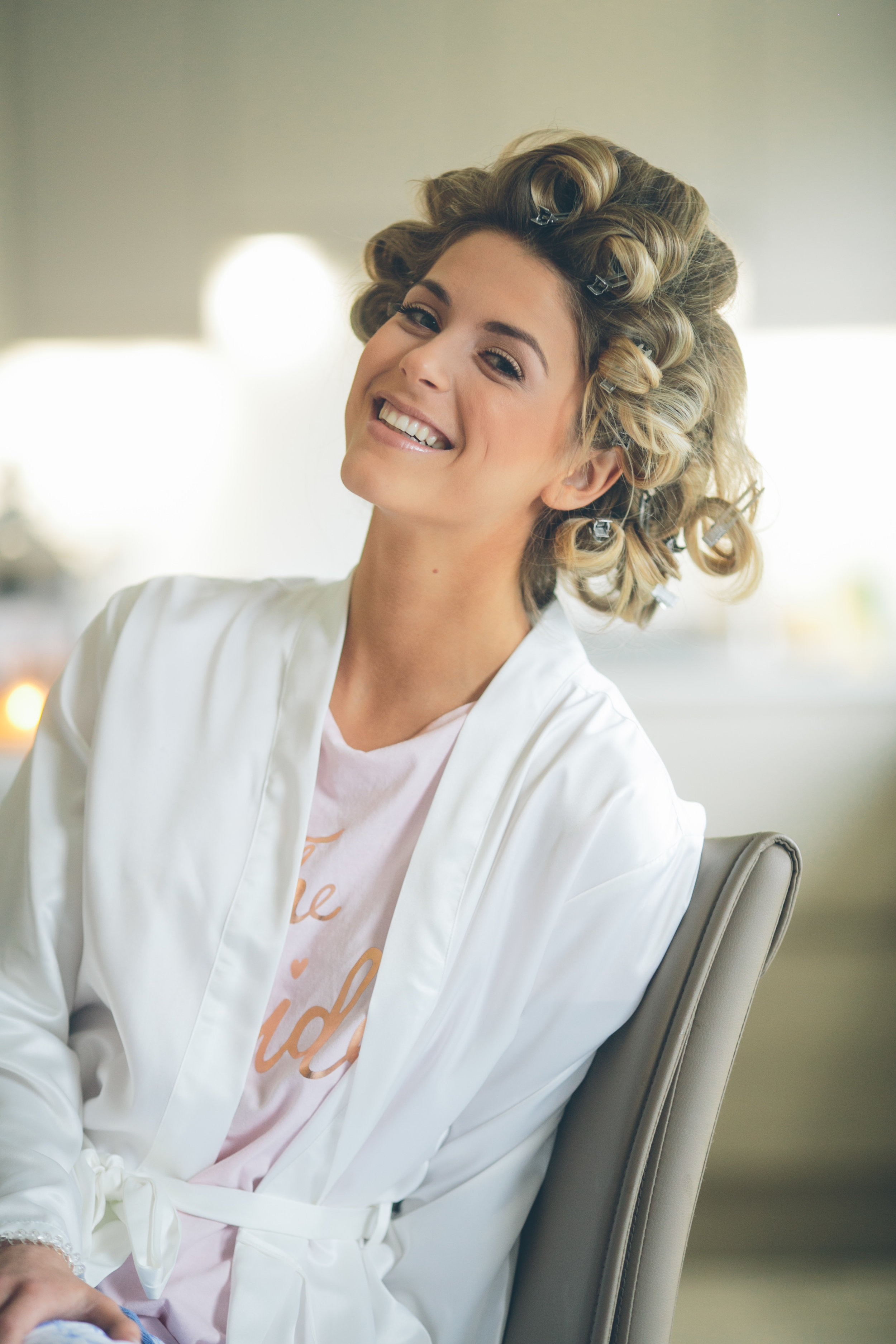 Woman smiling with hair rollers in her hair, wearing a white robe and a pink T-shirt, sitting on a chair in a room with blurred background.