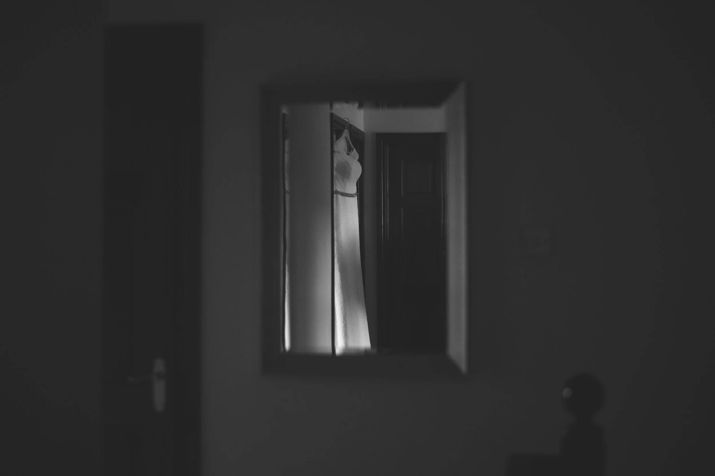 A black-and-white photo of a mirror hanging on a wall reflecting a wedding dress, with a dark door frame and a dresser with a rounded knob visible in the foreground.