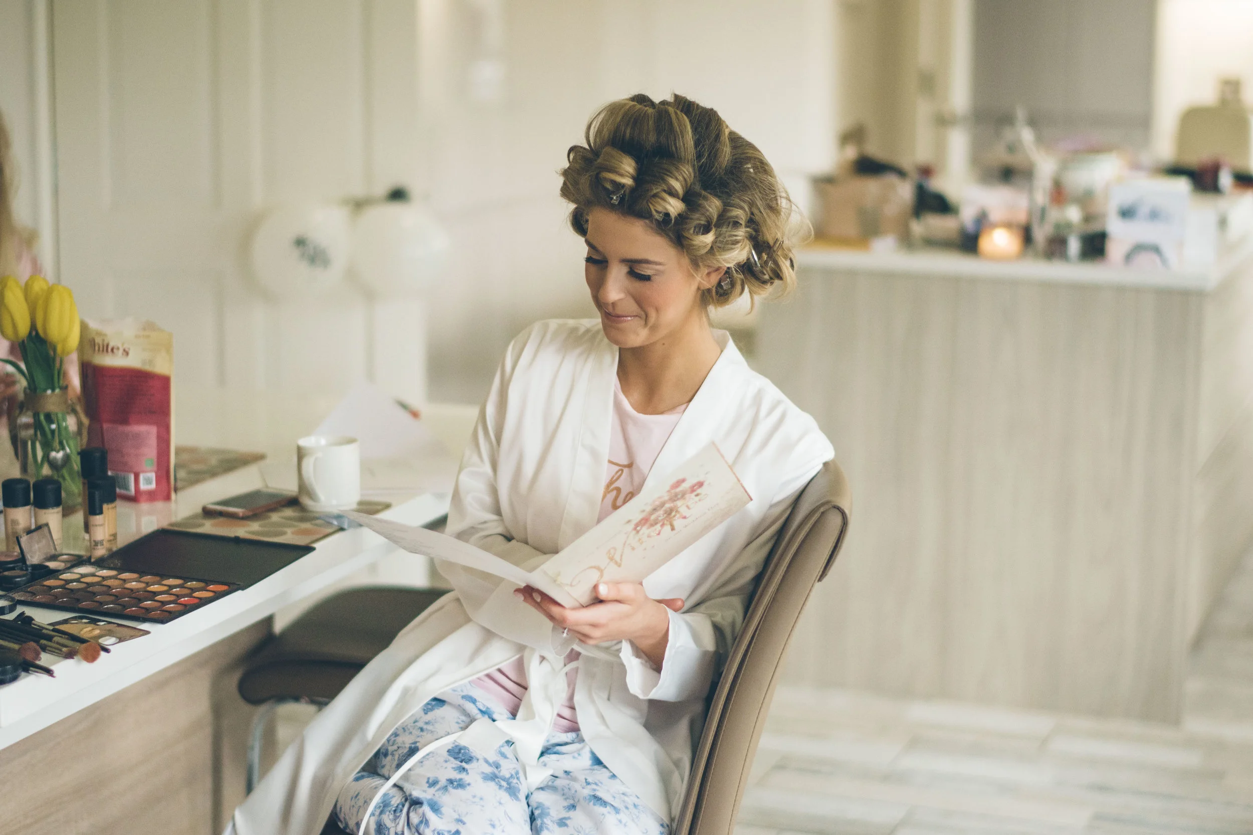 A woman with curled hair wearing a white robe, sitting at a vanity table reading a card.