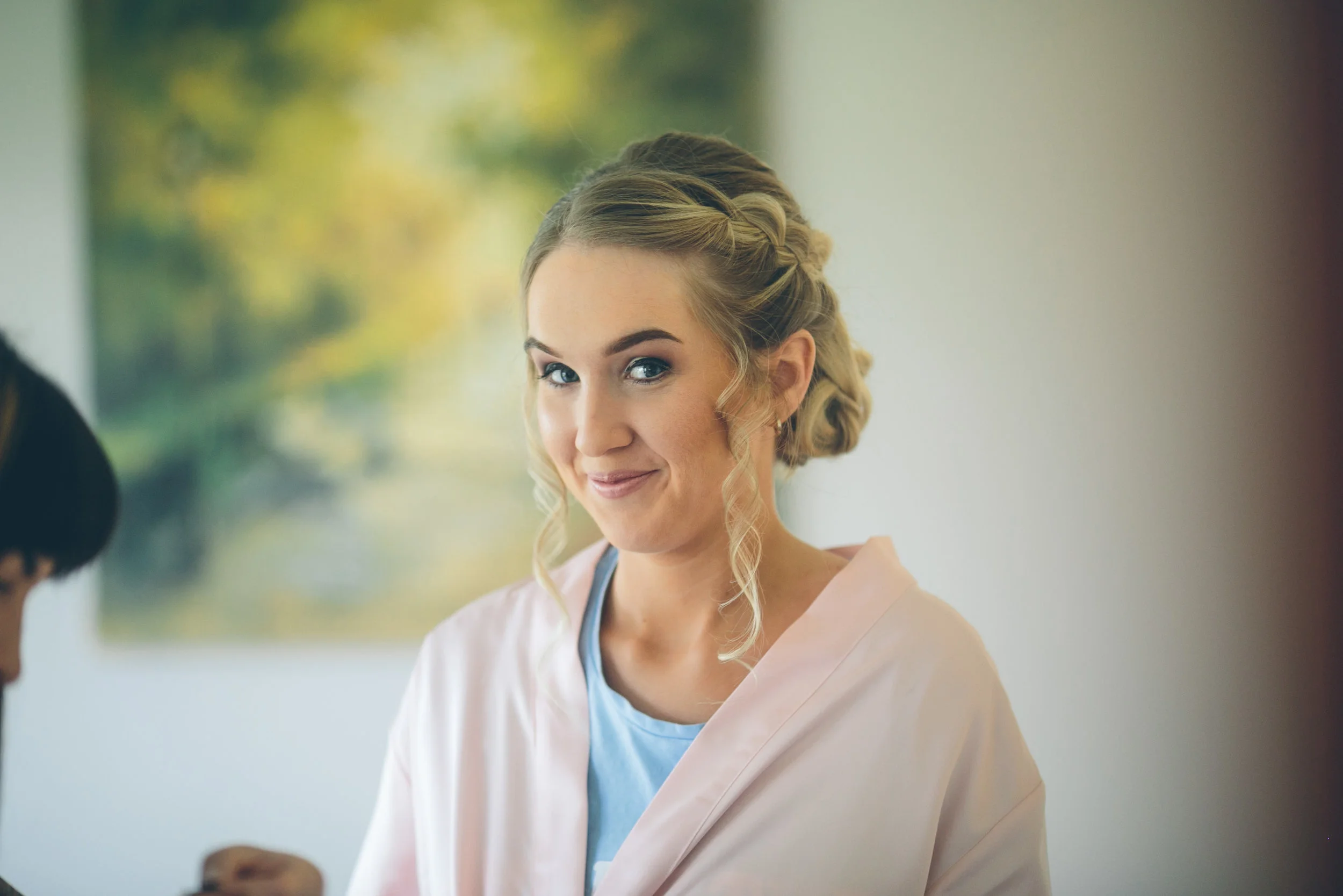 A young woman with blonde, curly hair styled in an elegant updo, smiling softly, wearing a light pink satin robe over a blue top. Background is softly blurred with outdoor greenery.