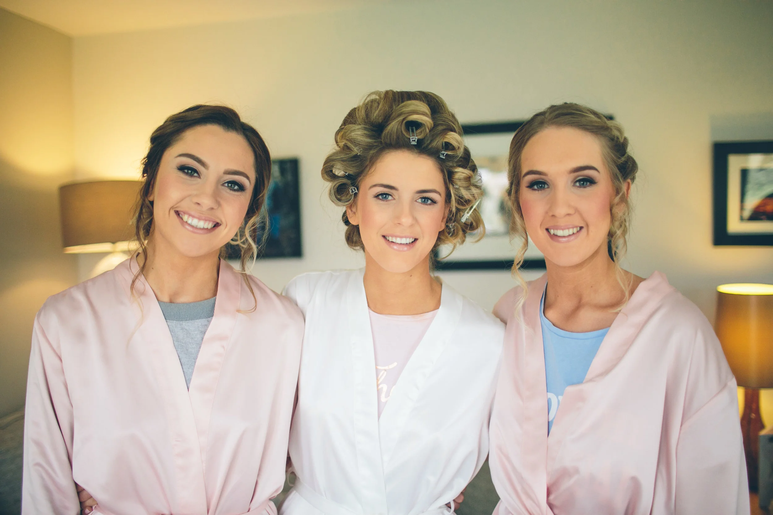 Three women in white and pink robes smiling, with one in curlers, in a room with lamps and framed pictures on the wall.