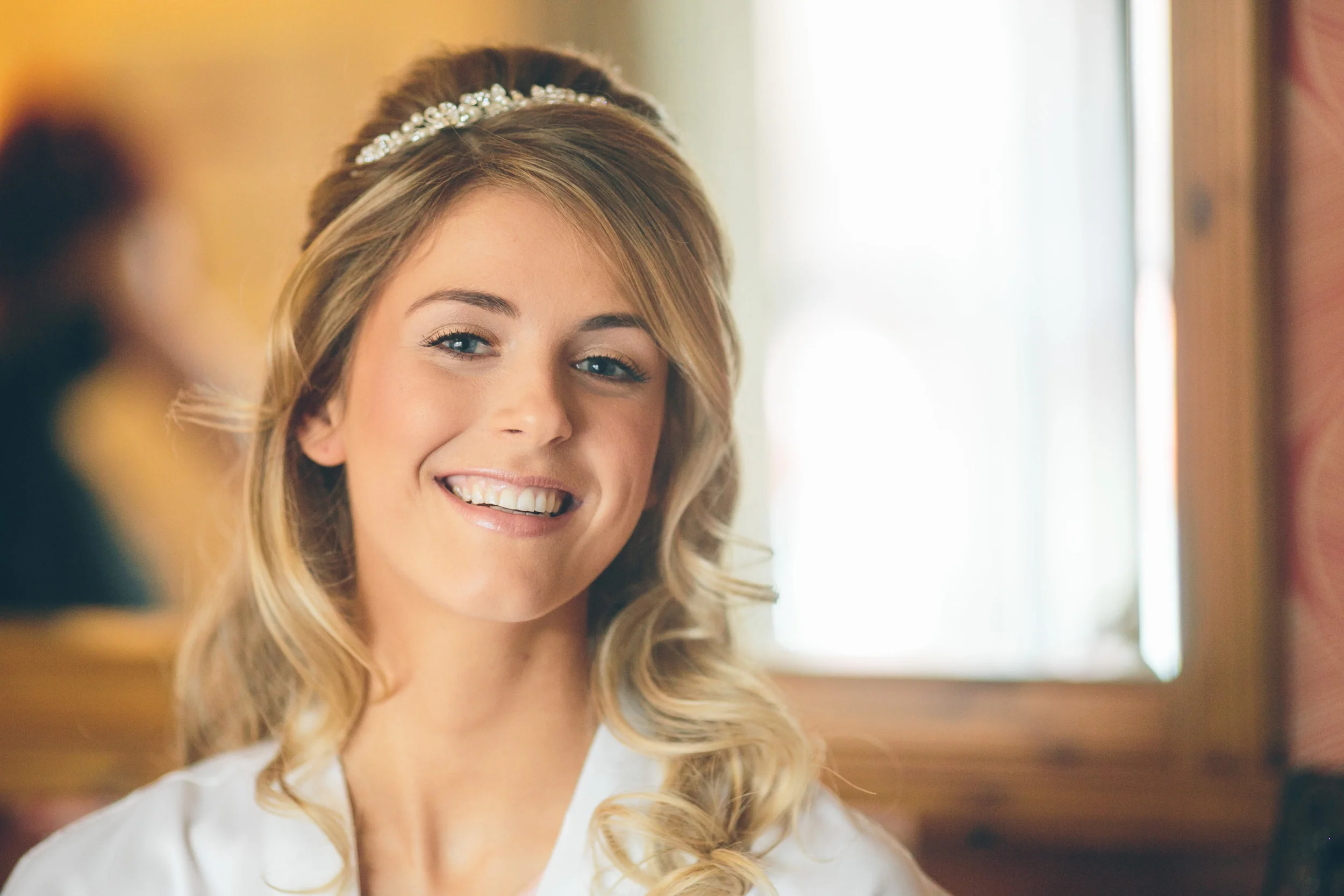 A smiling bride with wavy blonde hair, wearing a pearl headband, in a warmly lit room with a wooden window frame.