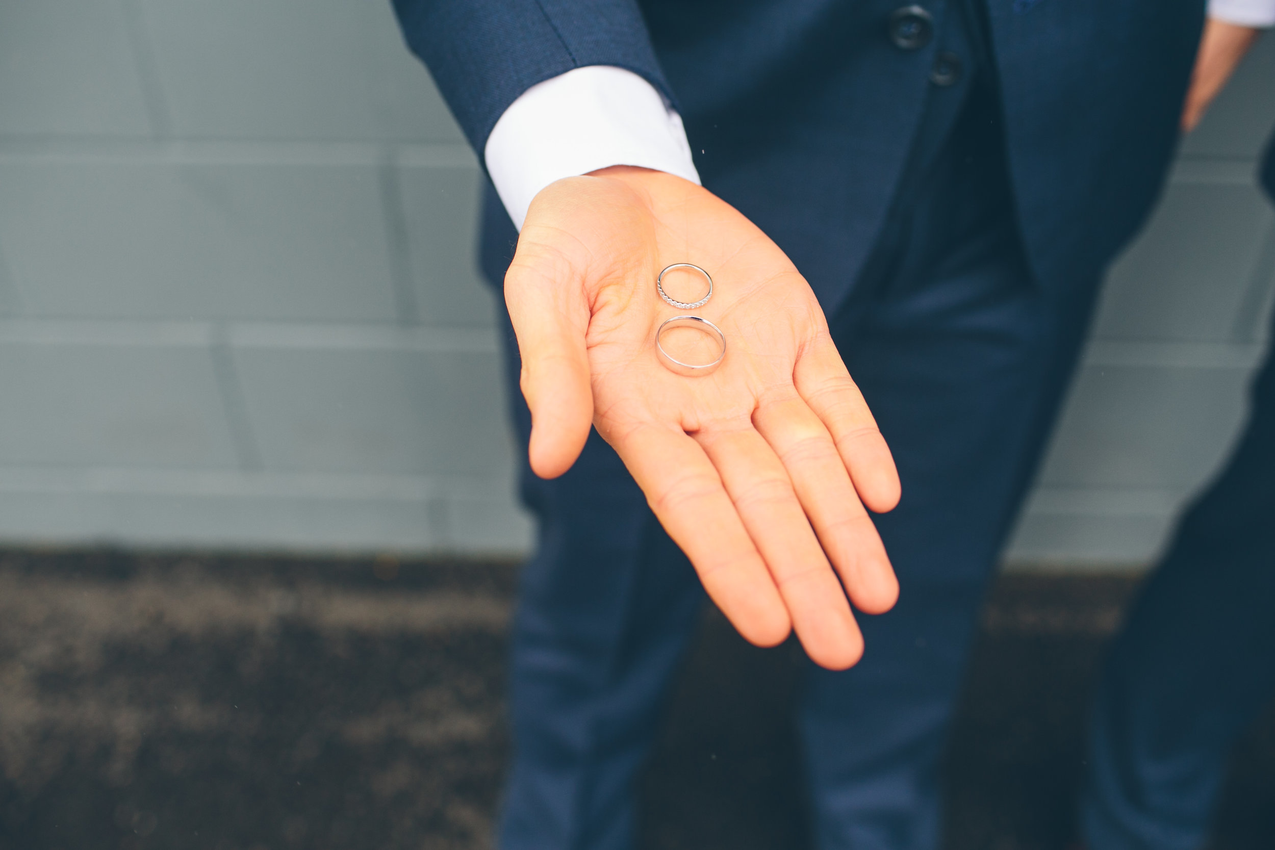 A person dressed in a dark suit is holding two wedding rings, one with a gemstone, in their open palm.