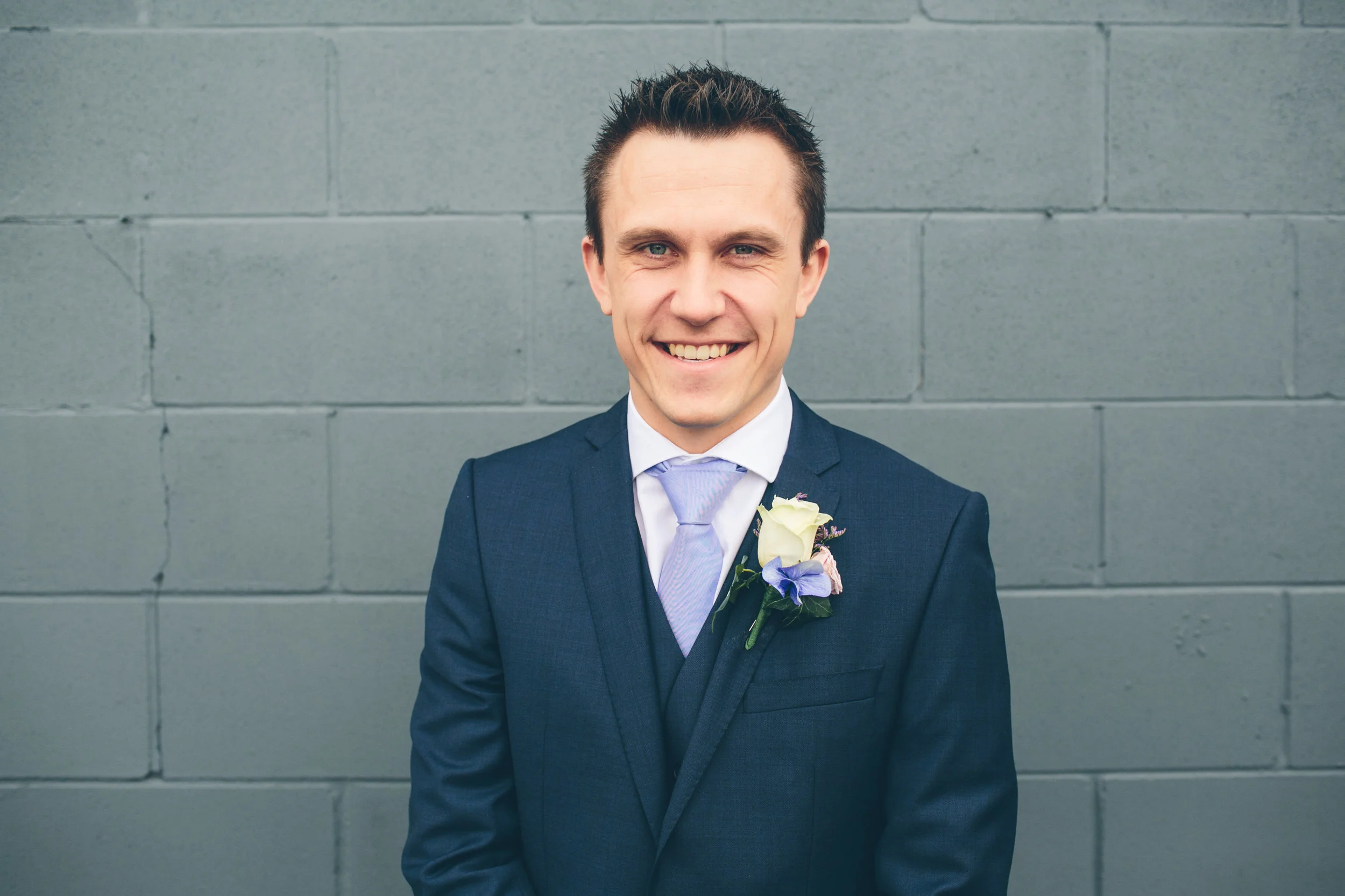 A smiling man in a blue suit with a light purple tie and a boutonnière stands against a gray brick wall.