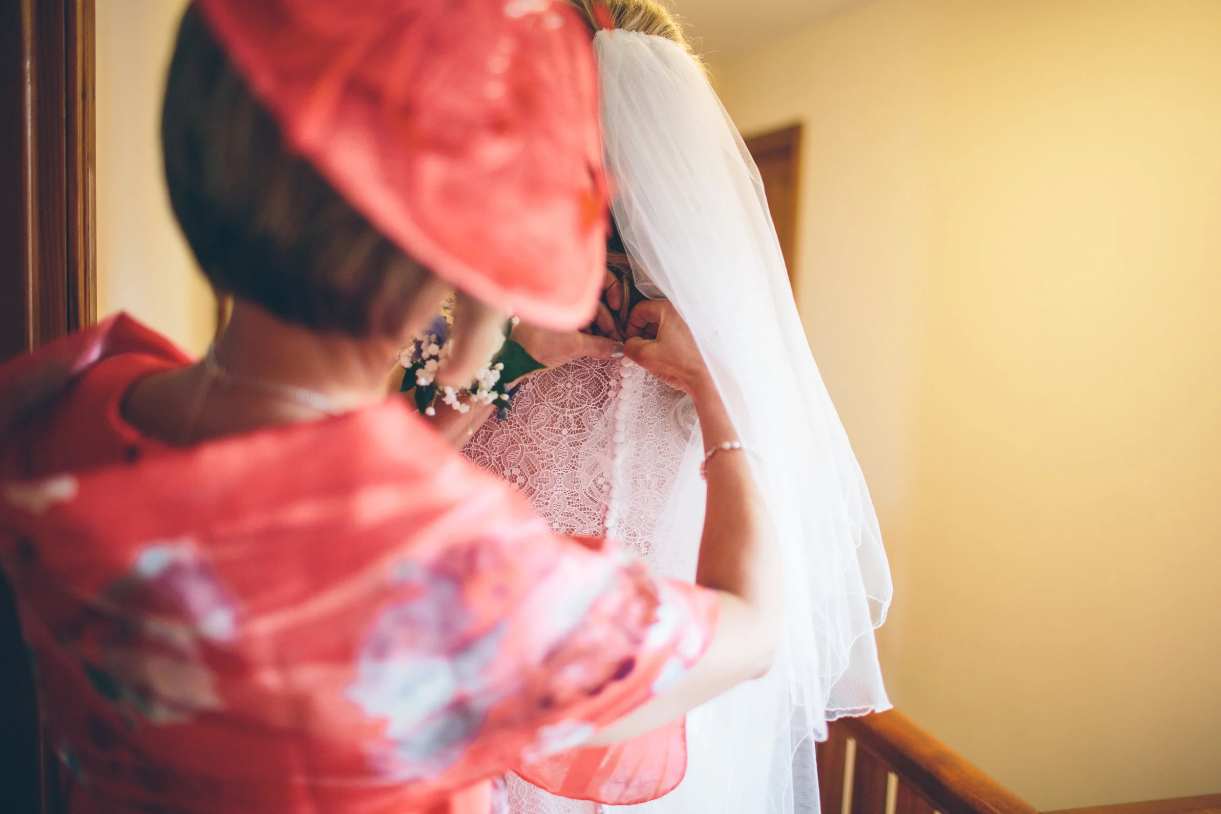 A woman in a pink floral dress and matching hat helps a bride with her wedding dress, which has lace details and a veil, as they prepare for the wedding inside a house or venue.