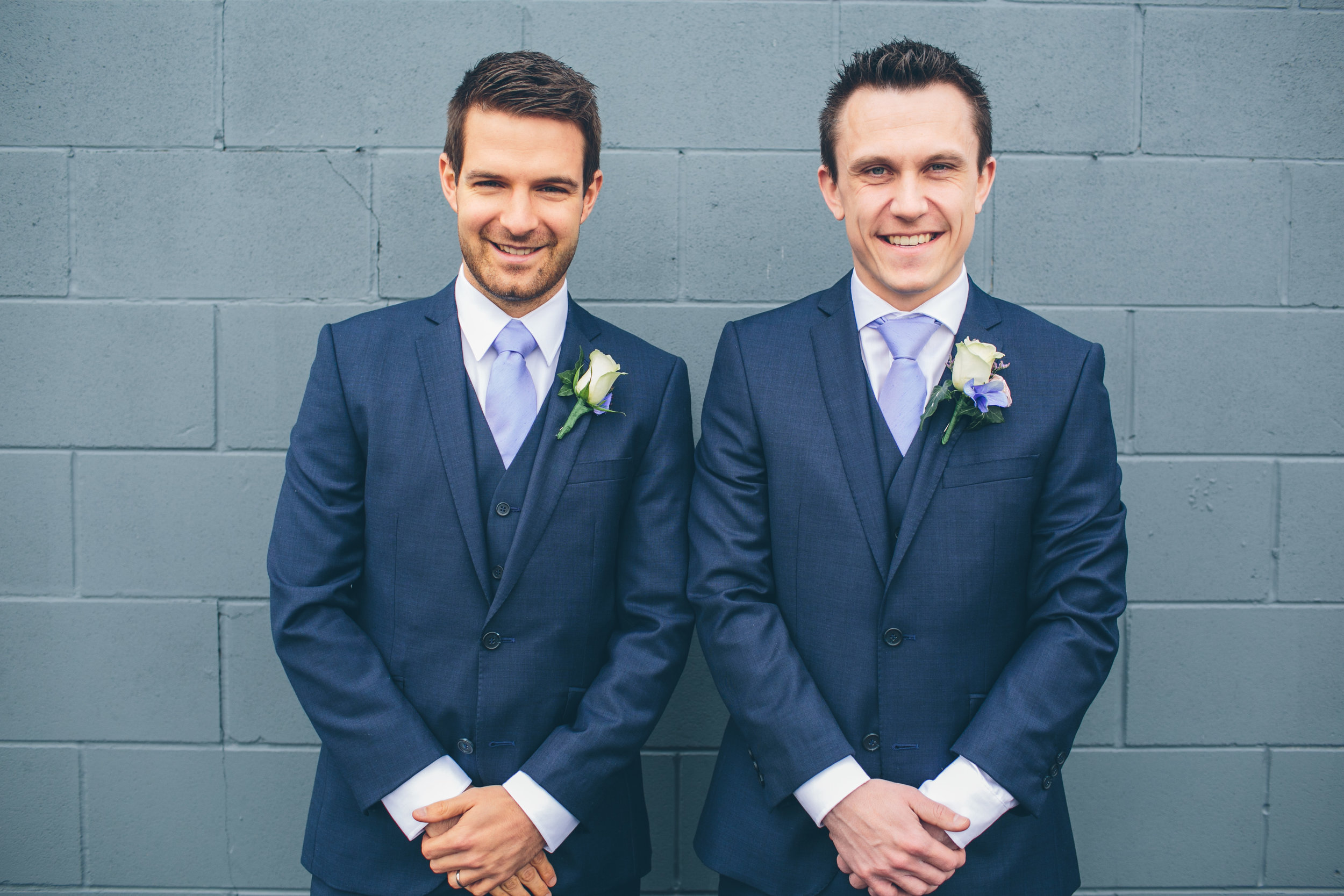 Two men in blue suits and light purple ties standing against a gray brick wall, both smiling, with white and purple boutonnieres on their lapels.
